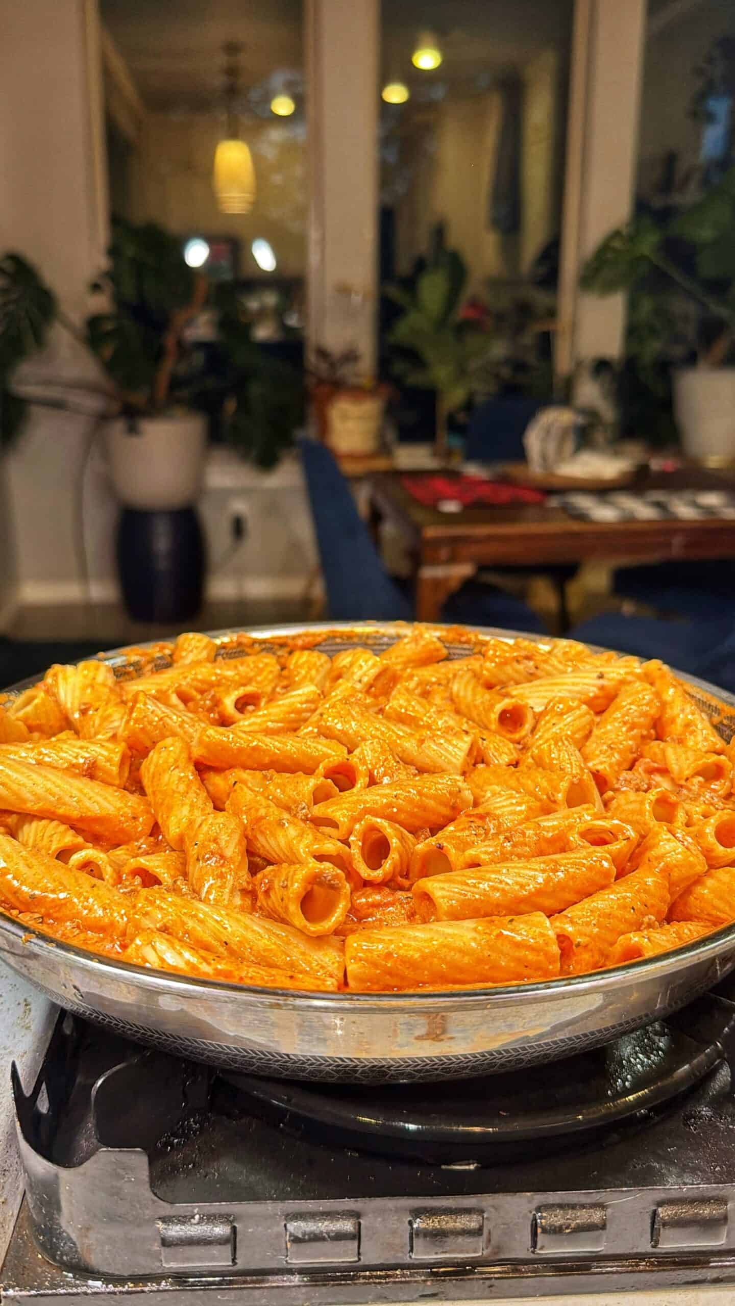 A skillet filled with rigatoni pasta in a red tomato sauce sits on a stovetop, with a cozy dining room and potted plants visible in the background.