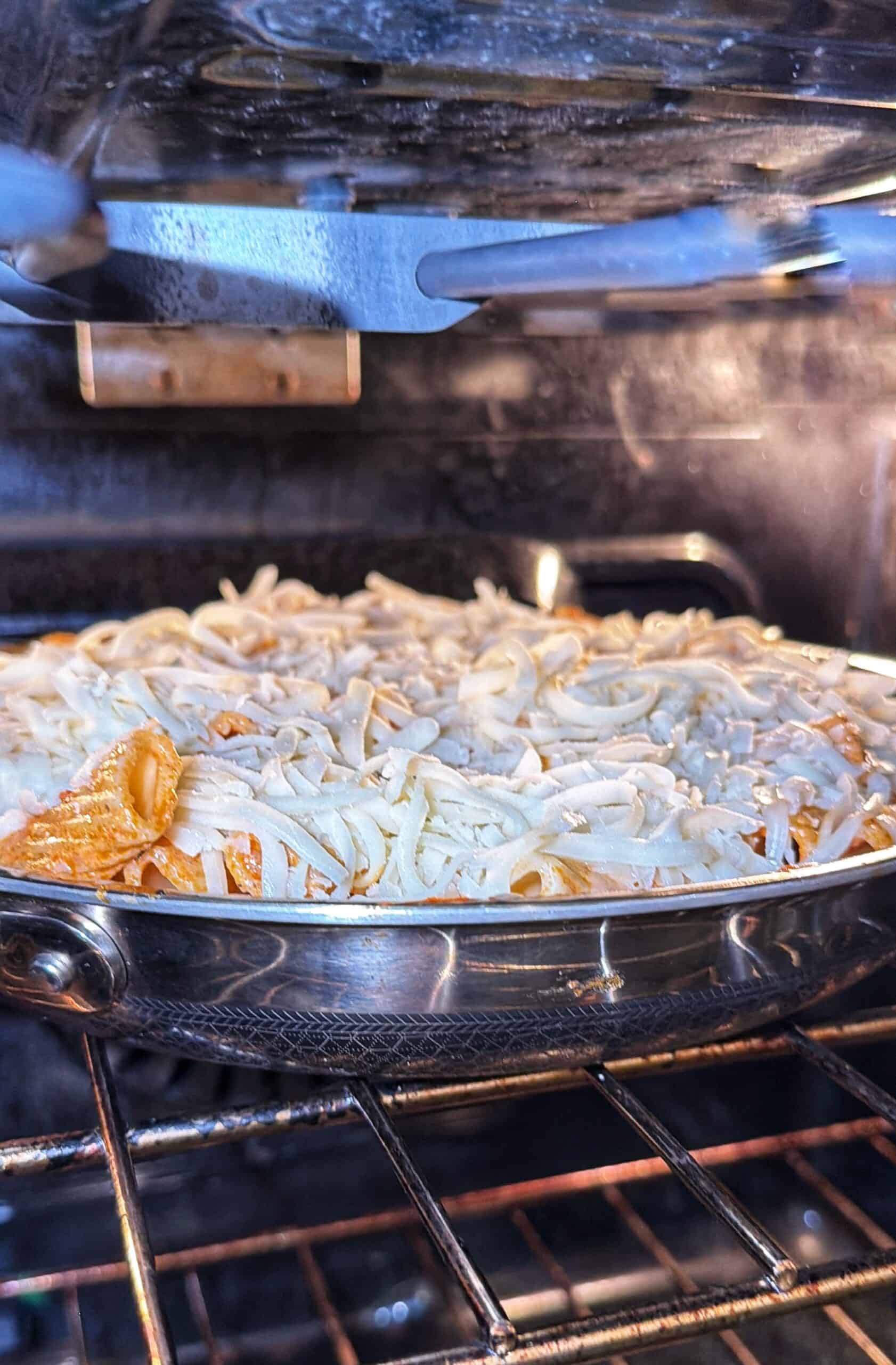 A metal pan filled with pasta covered in shredded cheese is baking on an oven rack. The oven is turned on, and the cheese is starting to melt.