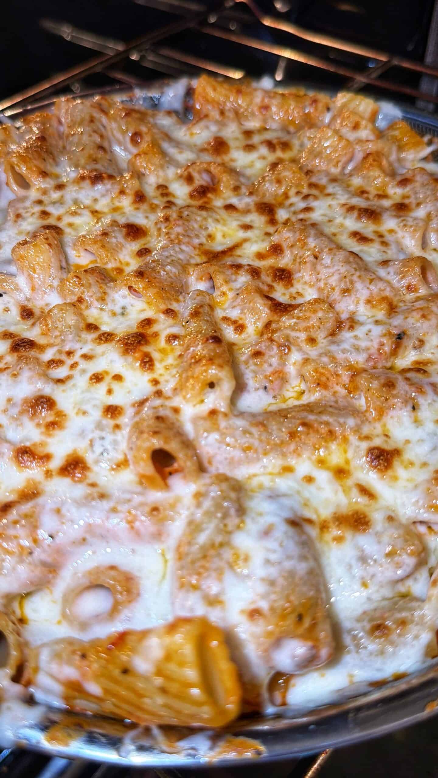 Baked pasta dish in an oven, topped with melted and lightly browned cheese, with rigatoni noodles visible beneath the cheese layer.