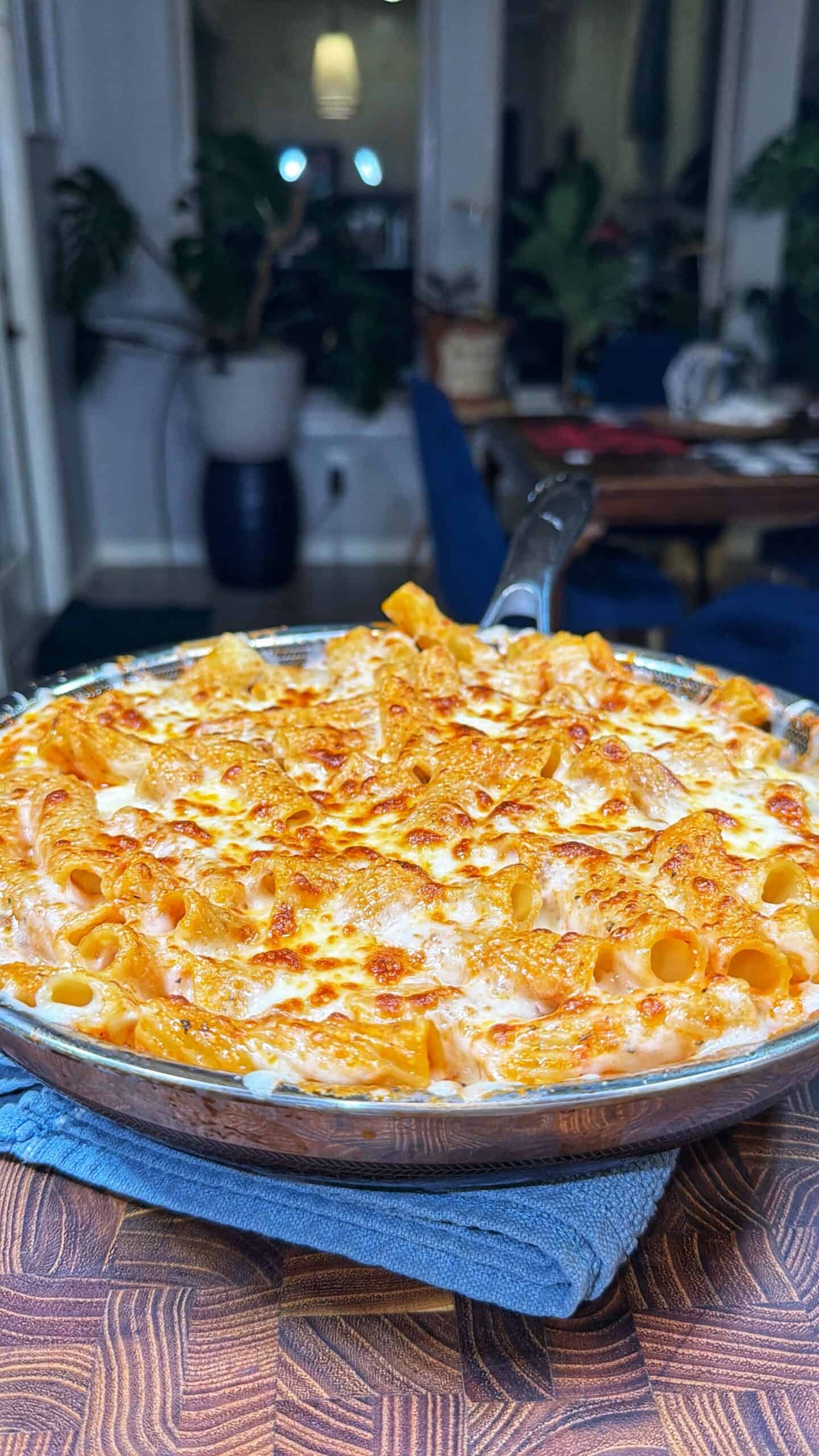 A cheesy baked pasta dish with rigatoni noodles is golden and bubbling in a round glass dish, resting on a blue cloth. The background shows a cozy, plant-filled dining room with a wooden table and blue chairs.