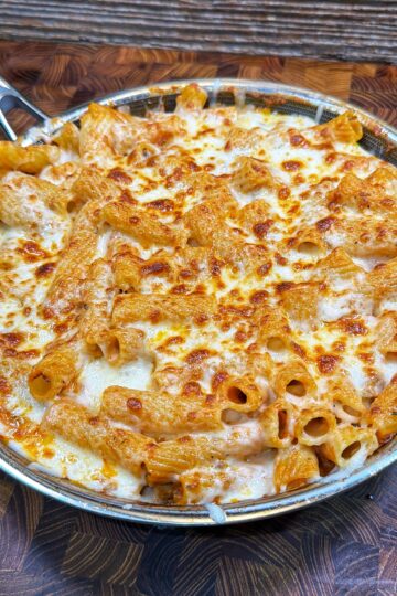 A skillet filled with baked pasta, topped with melted and browned cheese, sits on a wooden surface. The pasta appears to be rigatoni, coated in a creamy, tomato-based sauce.