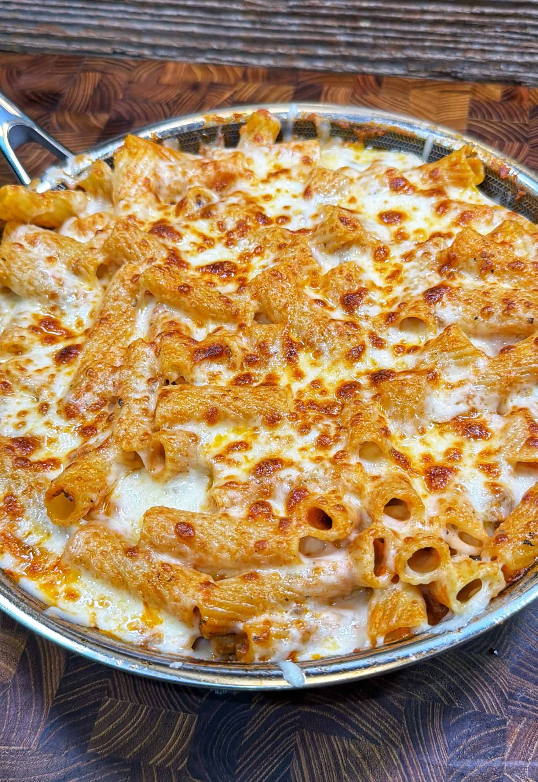 A skillet filled with baked pasta, topped with melted and browned cheese, sits on a wooden surface. The pasta appears to be rigatoni, coated in a creamy, tomato-based sauce.