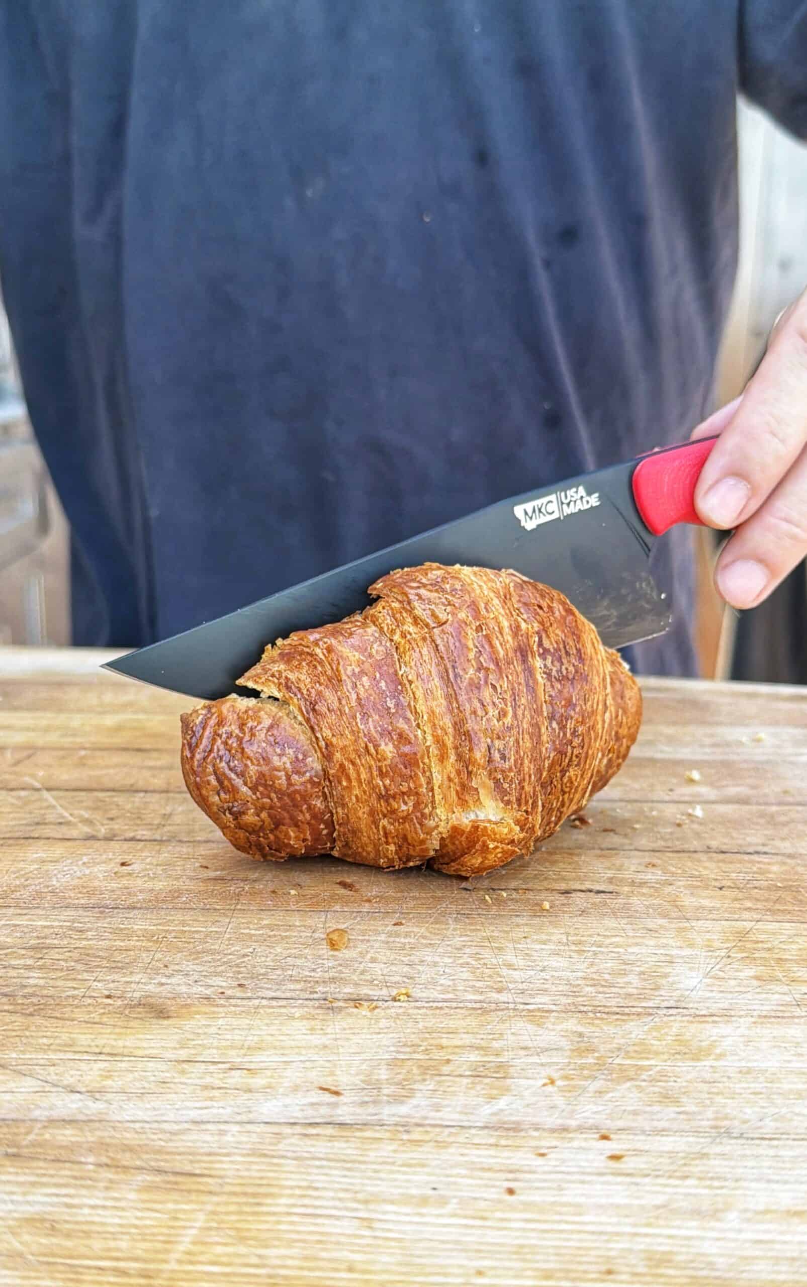 A person cuts a golden-brown croissant on a wooden board with a black and red-handled knife, wearing a dark shirt.