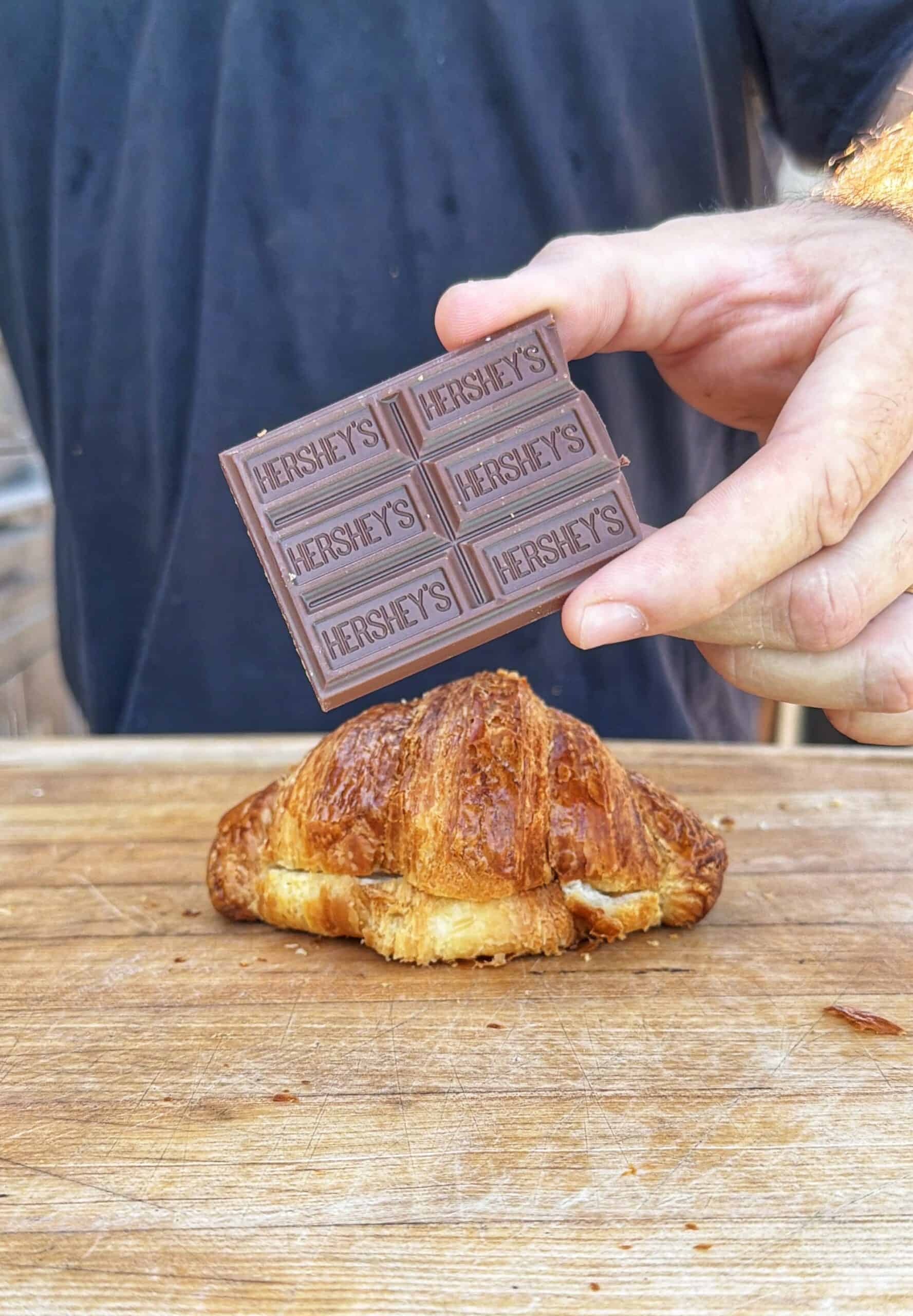 A person holds a Hersheys chocolate bar above a croissant on a wooden surface, with the persons torso visible in the background.