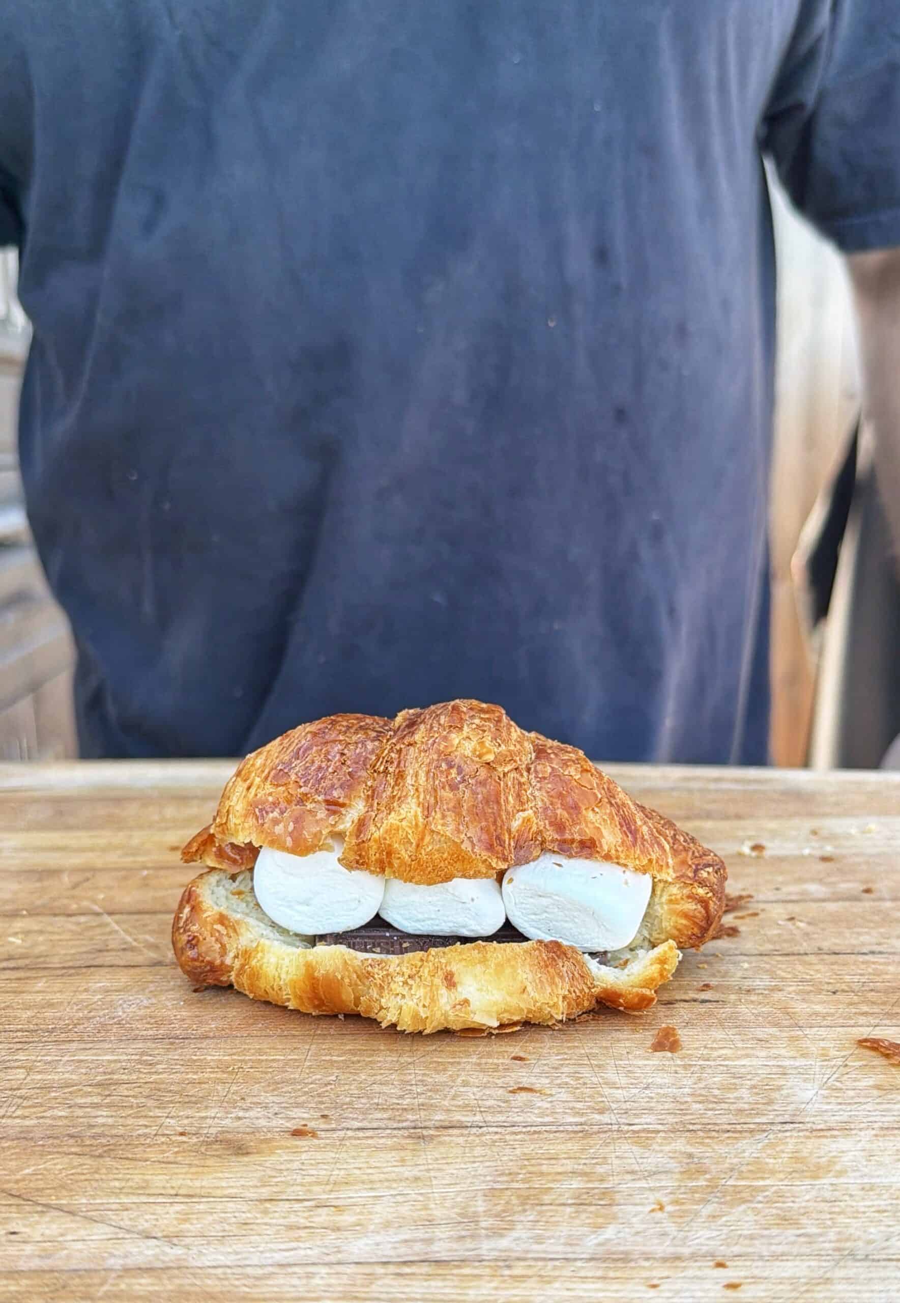A croissant filled with three marshmallows and a piece of chocolate sits on a wooden surface, with a person in a dark shirt standing in the background.
