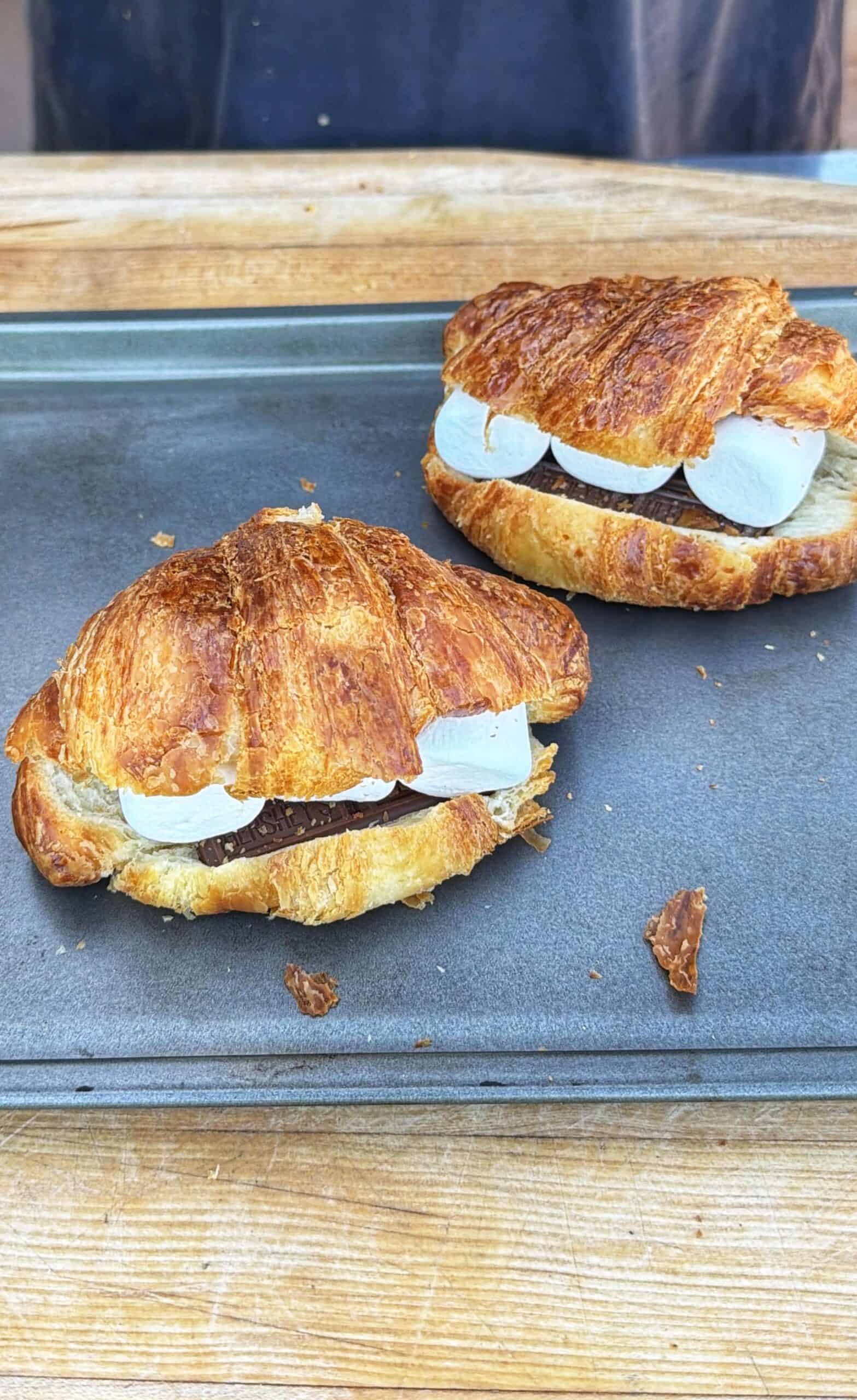 Two croissants filled with chocolate and marshmallows are placed on a baking tray atop a wooden surface.