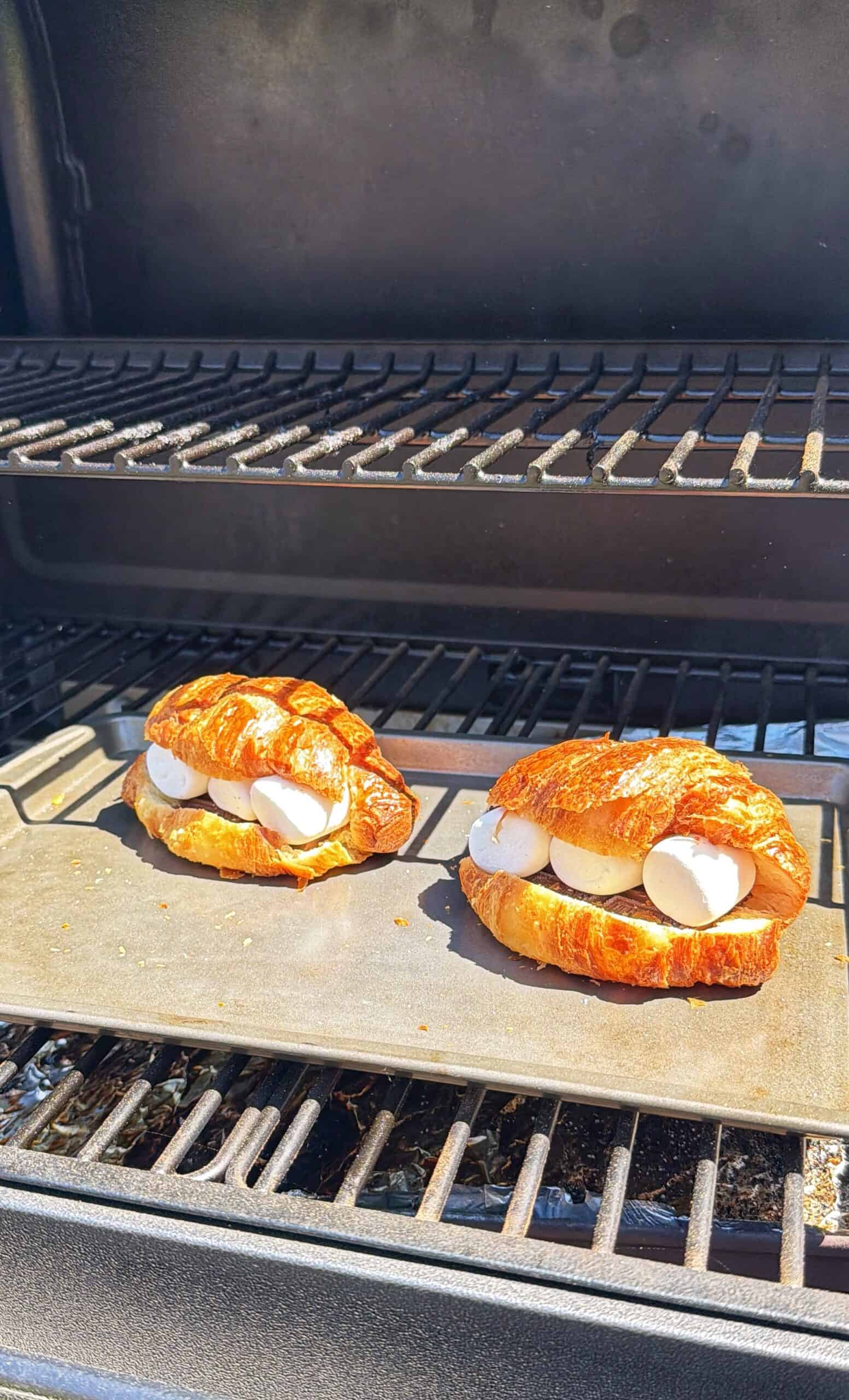 Two croissants filled with marshmallows are placed on a baking tray inside a grill, ready to be toasted. Sunlight shines on the food, giving the scene a warm, outdoor feel.