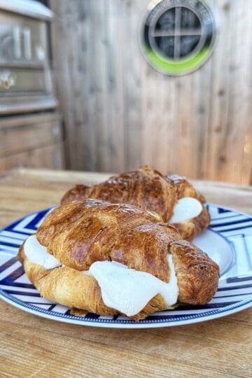 Two croissants filled with white cheese are placed on a blue and white patterned plate, resting on a wooden table outdoors with a wooden fence and grill in the background.