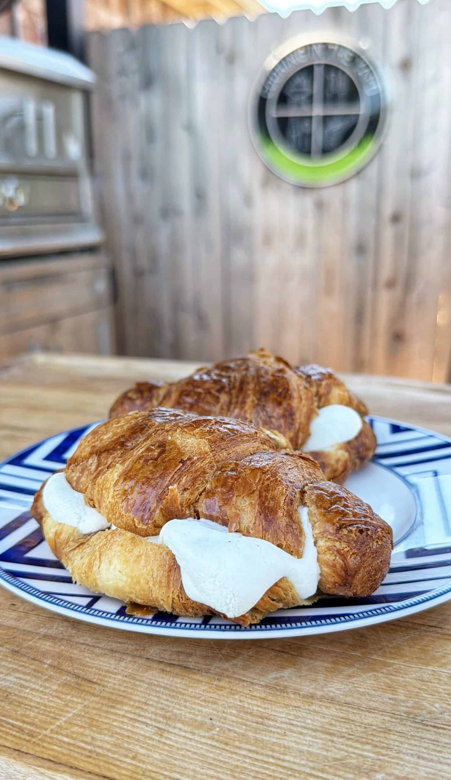 Two croissants filled with white cheese are placed on a blue and white patterned plate, resting on a wooden table outdoors with a wooden fence and grill in the background.