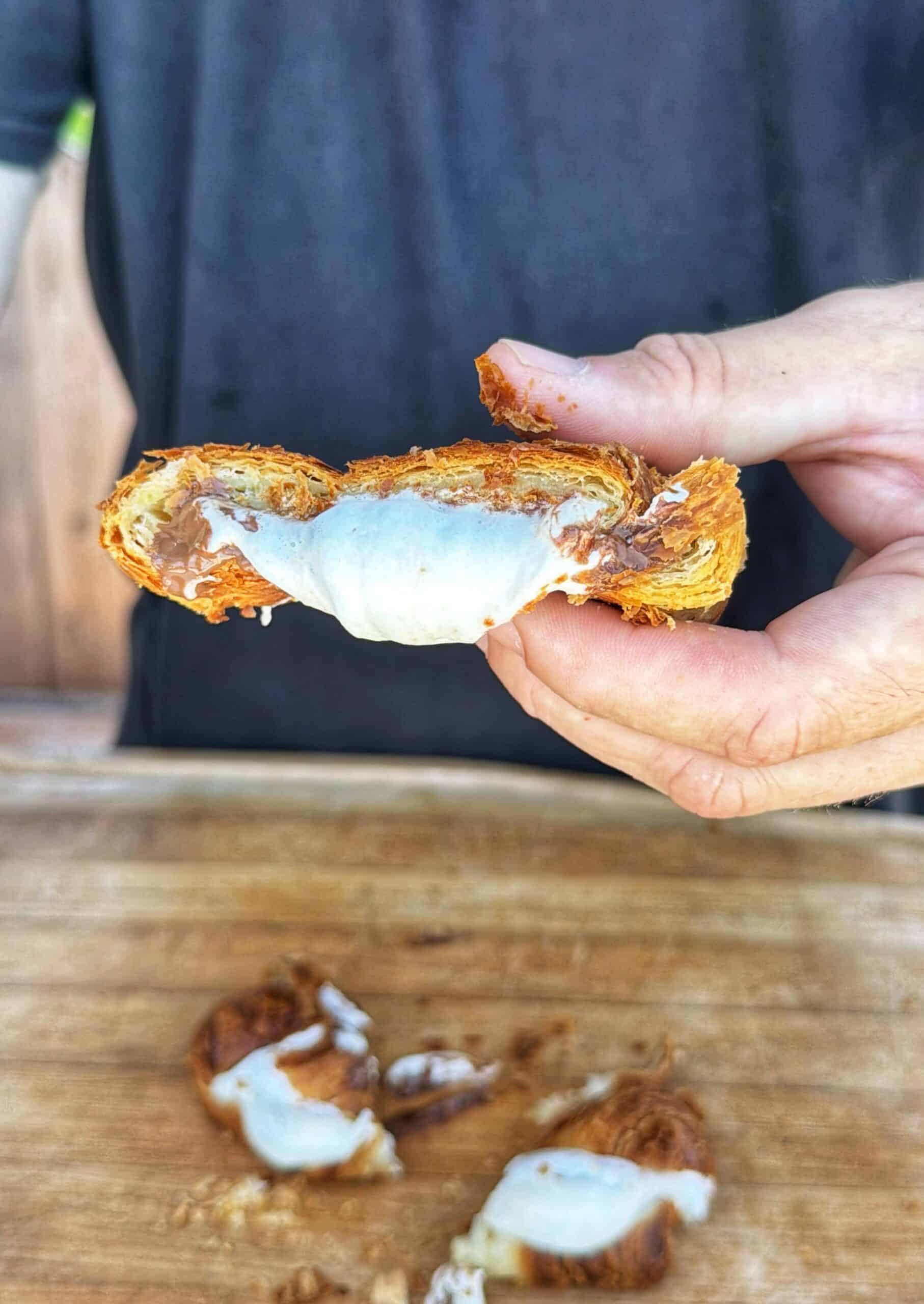 A person holds a halved croissant showing a gooey chocolate and cream filling, with more pieces of croissant on a wooden surface below.