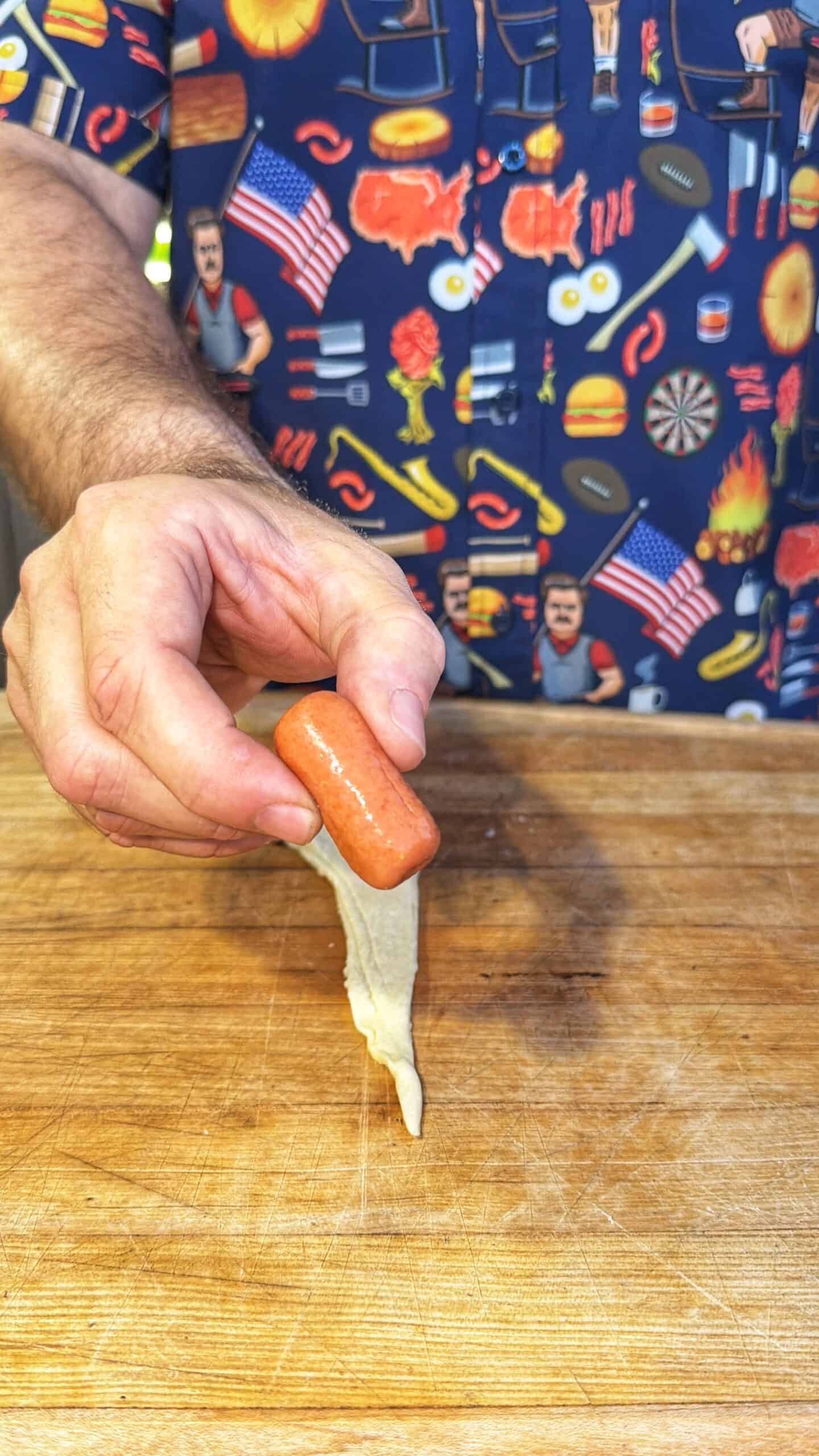A person in a colorful, food-themed shirt holds a small sausage over a strip of raw dough on a wooden surface, preparing to wrap the sausage.