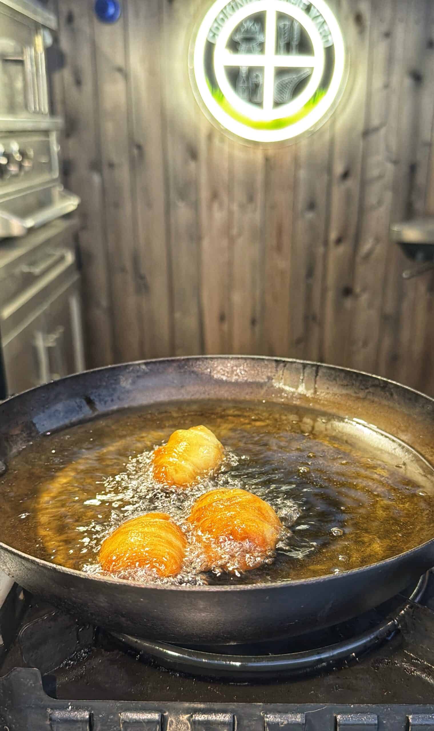 Three pieces of dough are frying in hot oil in a black pan on a stove, with bubbles forming around them. The kitchen has wooden walls and a round window with sunlight shining through.