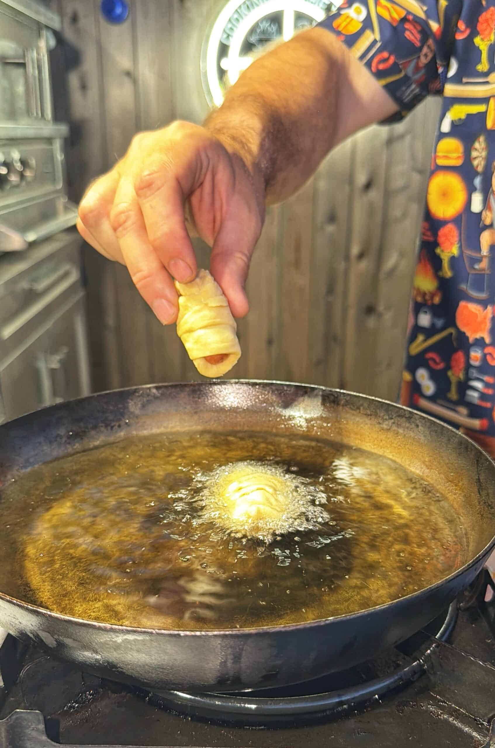 A person’s hand lowers a rolled pastry into hot oil in a skillet on a stove, causing bubbles to form. The background shows a wooden wall and part of a patterned shirt sleeve.