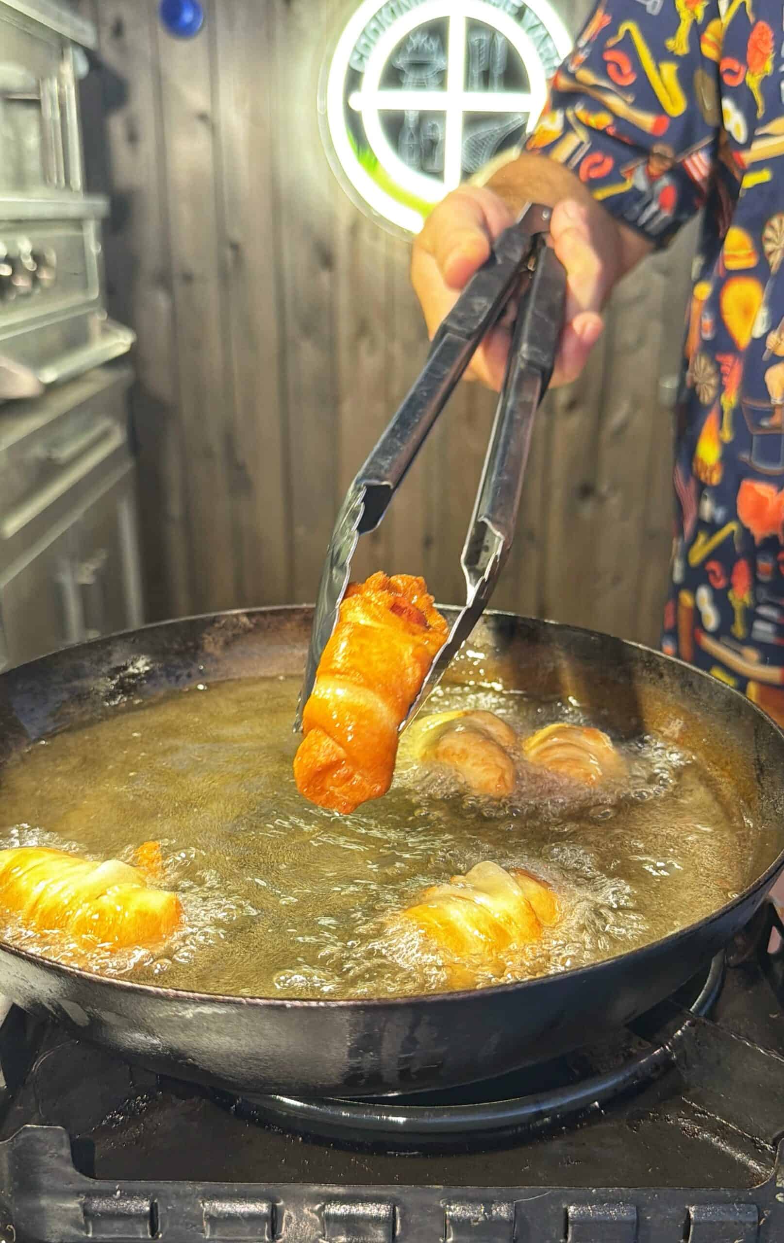 A person uses tongs to hold a golden-brown fried food roll above a hot skillet filled with oil, with more rolls frying in the pan. The kitchen background includes a window and metal appliances.