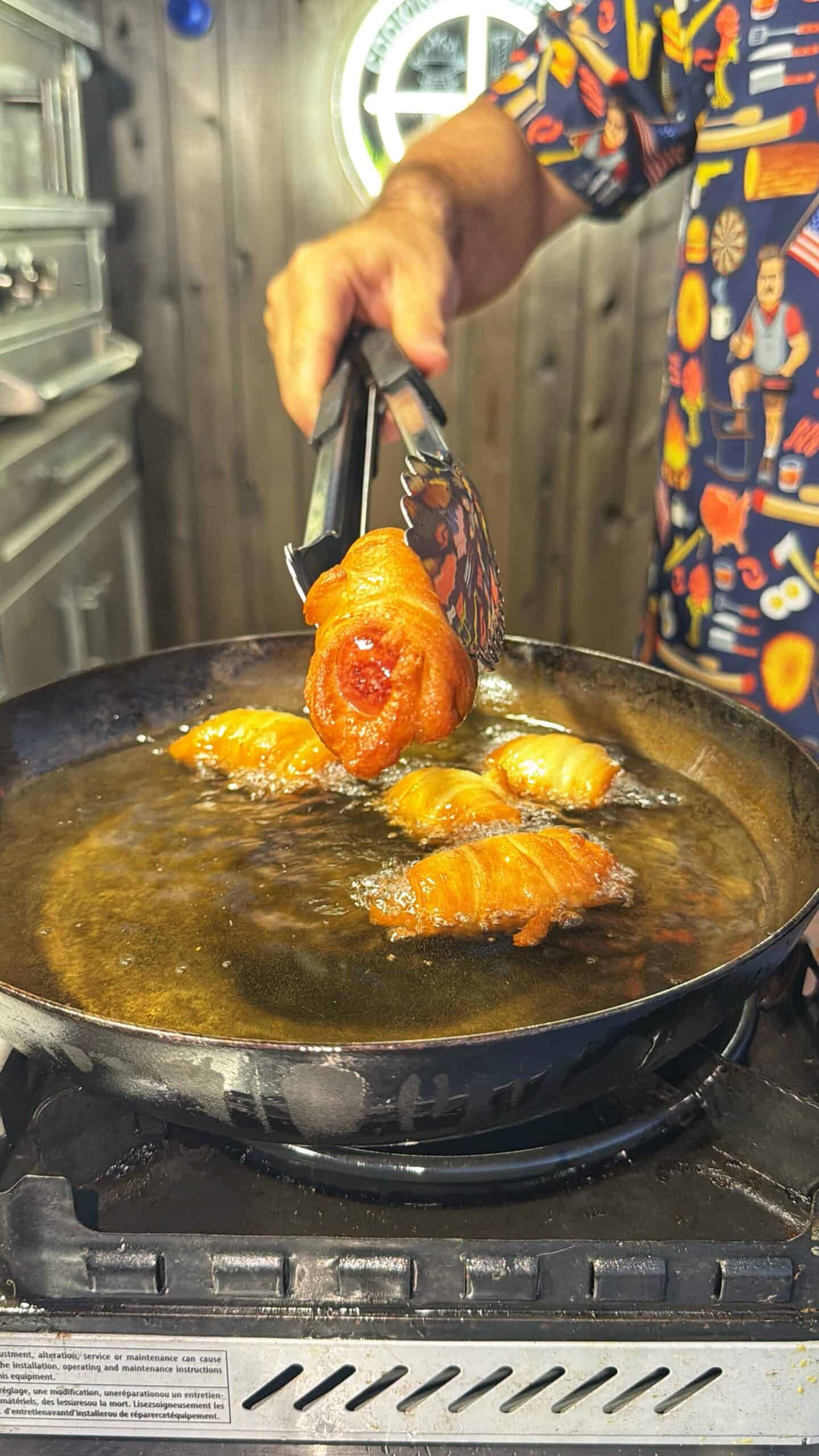 A person using tongs to fry bacon-wrapped food in a pan of oil on a stovetop, wearing a colorful shirt with food patterns. The background is a wooden kitchen wall.