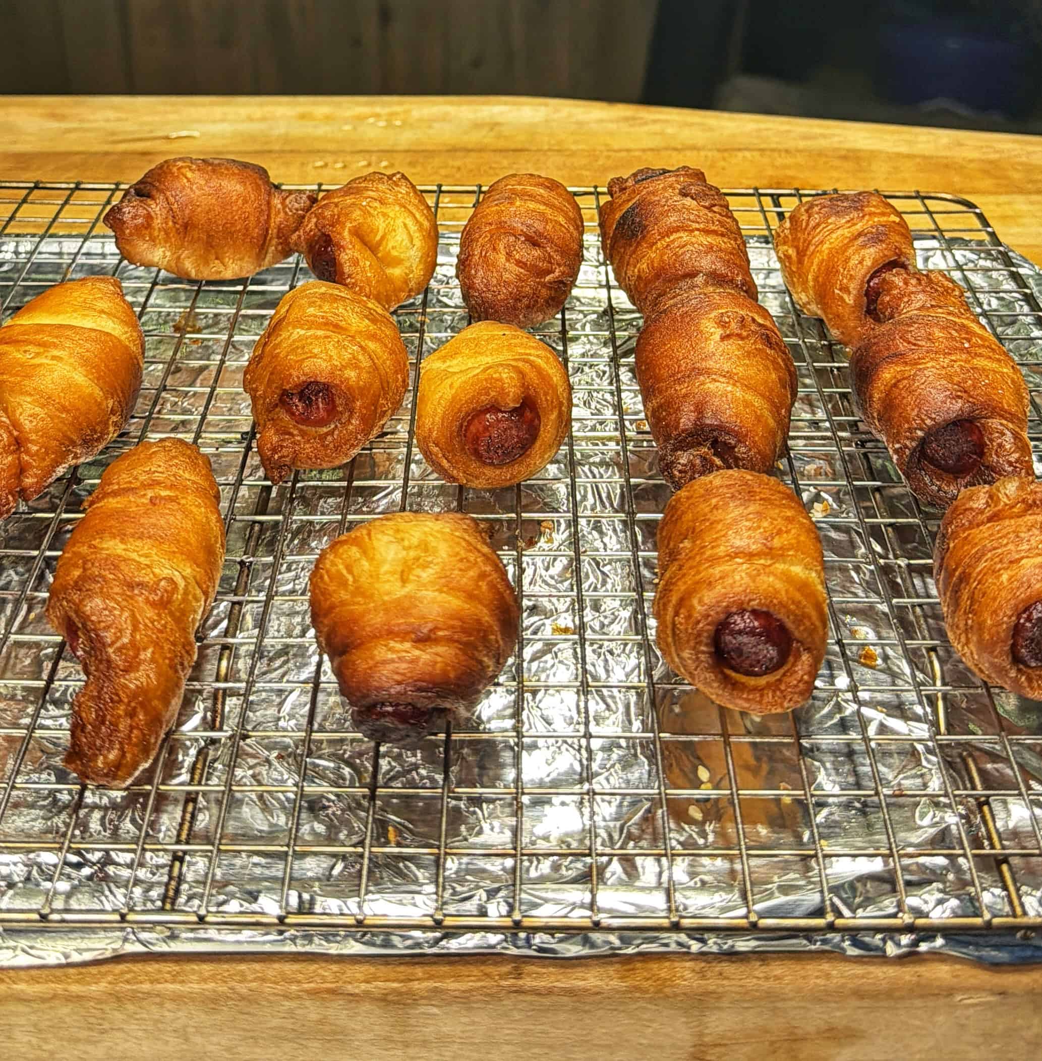 Golden-brown croissant rolls cooling on a wire rack lined with foil atop a wooden surface. The pastries appear flaky and crisp, arranged in rows.