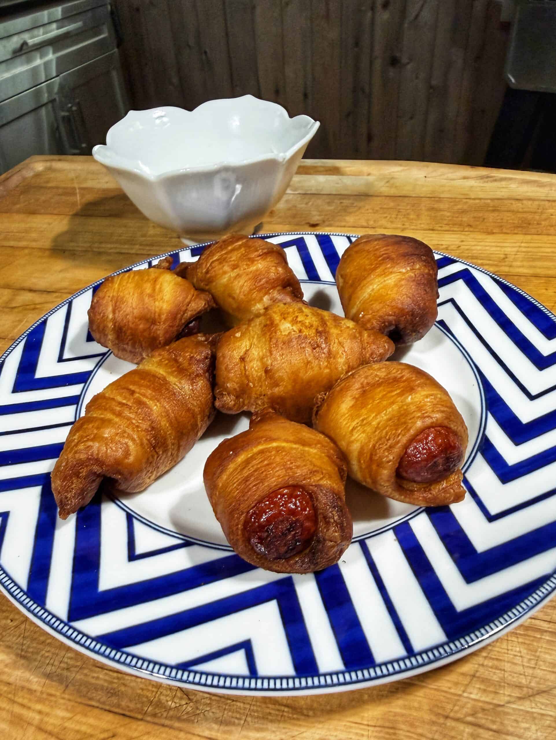 A plate with six golden-brown crescent rolls, some with sausages inside, sits on a wooden counter. A white bowl is in the background. The plate has a blue and white chevron pattern.