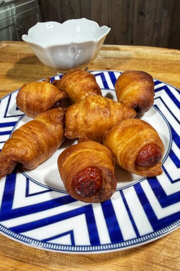 A plate with six golden-brown pigs in a blanket is set on a blue and white patterned dish. A white bowl sits in the background on a wooden surface.
