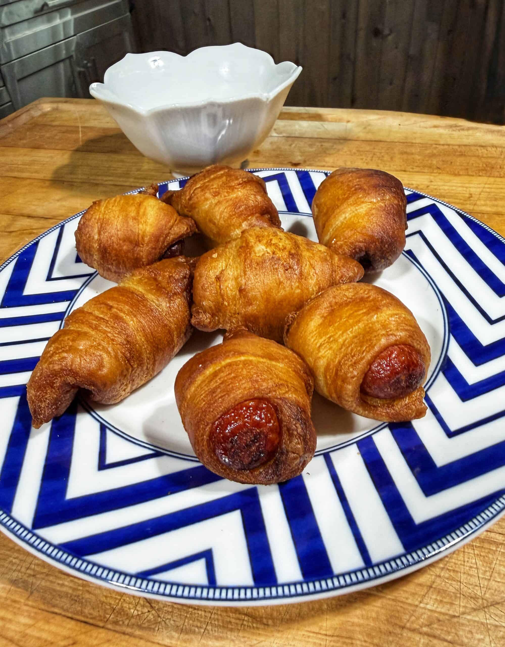 A plate with six golden-brown pigs in a blanket is set on a blue and white patterned dish. A white bowl sits in the background on a wooden surface.