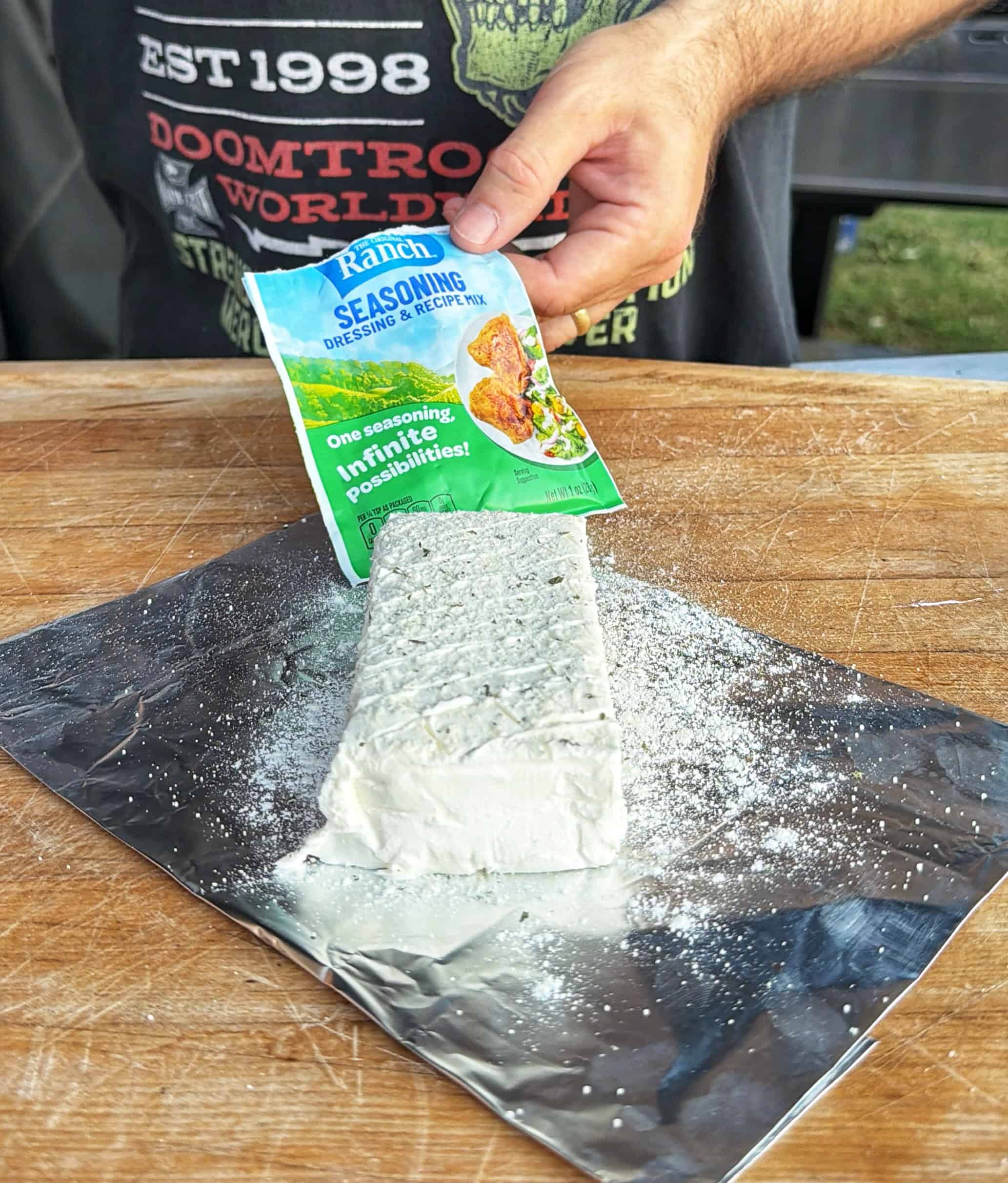 A person sprinkles Ranch seasoning mix over a block of cream cheese placed on a sheet of aluminum foil on a wooden surface.