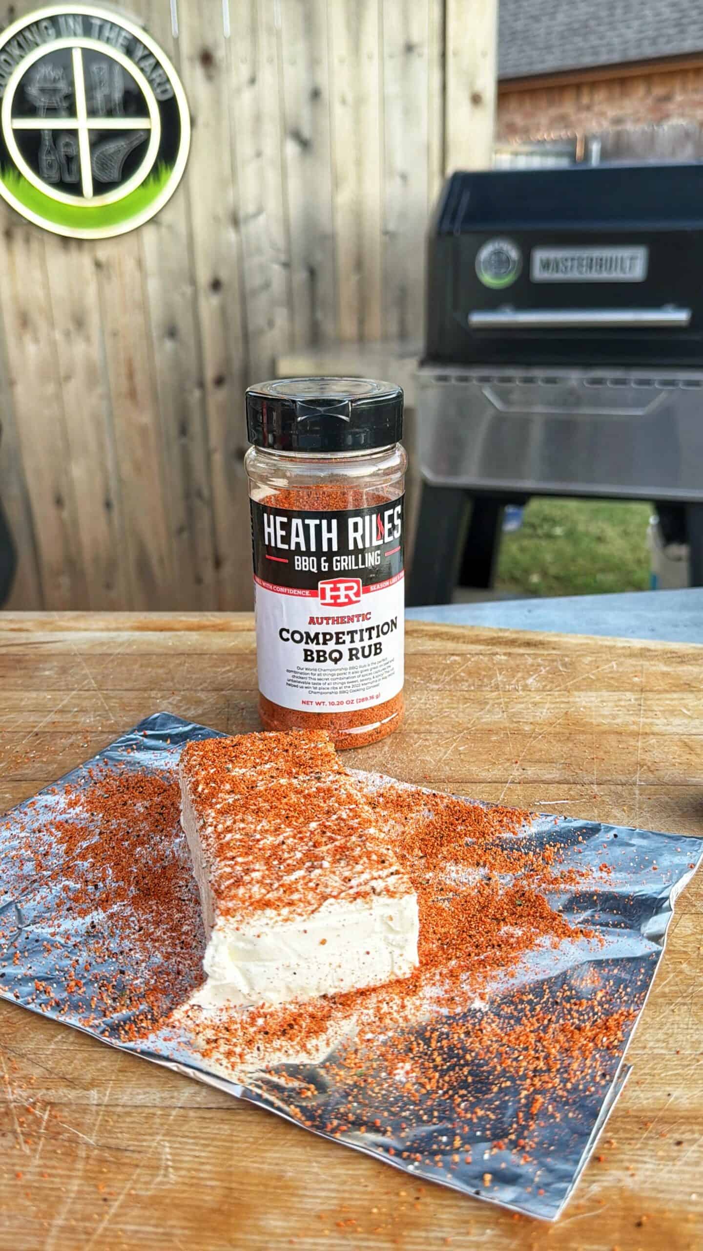 A block of cream cheese covered with orange BBQ rub sits on foil next to a Heath Riles BBQ & Grilling Competition BBQ Rub container, with a grill and wooden fence in the background.
