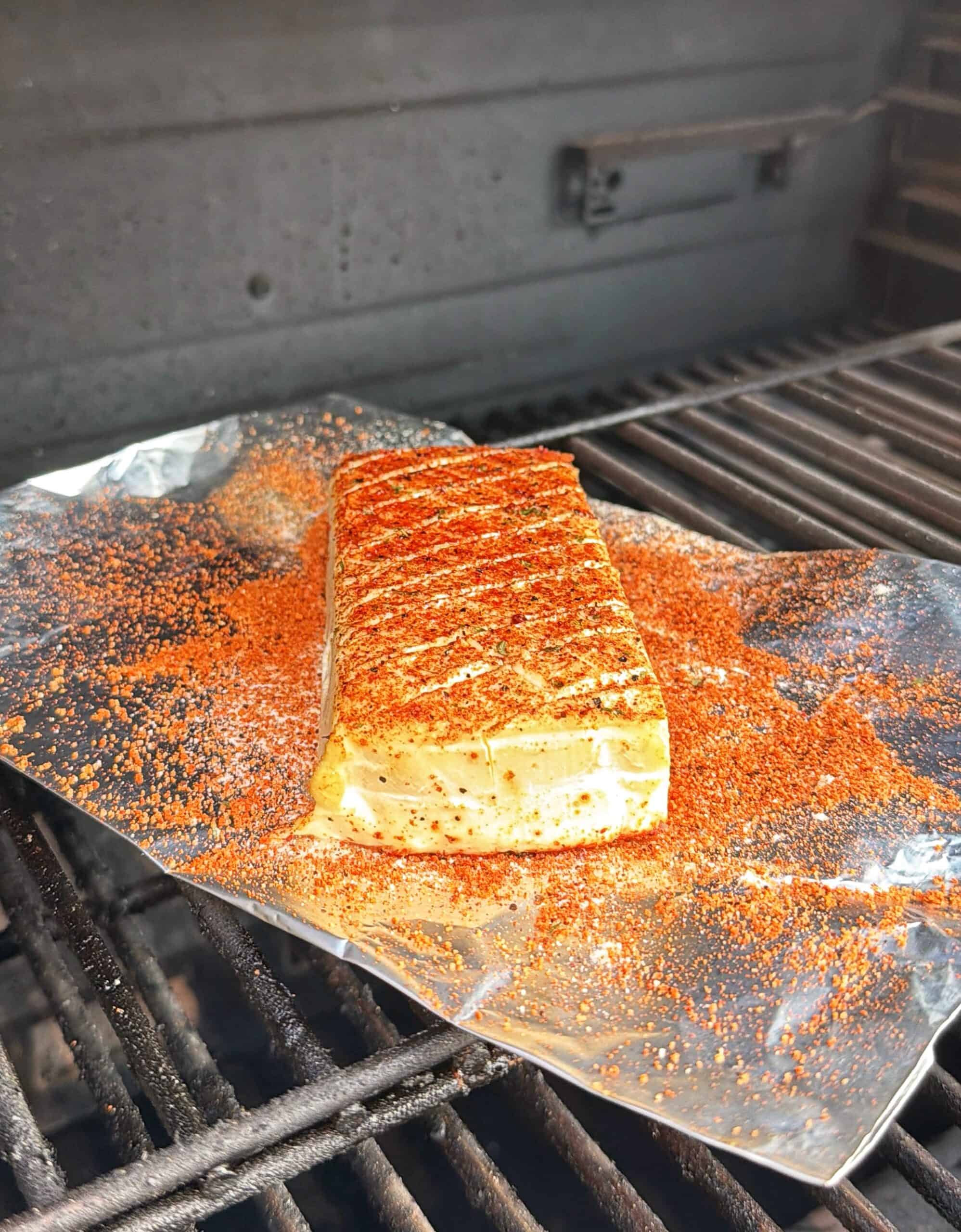 A block of seasoned cheese sits on a sheet of foil atop a grill, dusted with red spices and grill marks visible on top, inside an open barbecue grill.