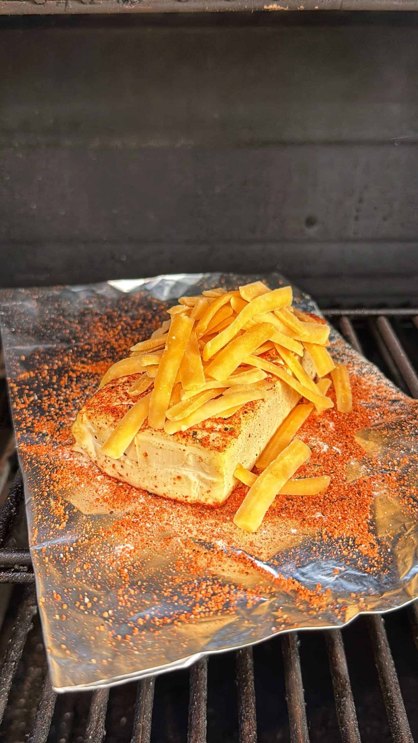 A block of seasoned cheese on a piece of foil sits on a grill, topped with crispy tortilla strips and sprinkled with spice.