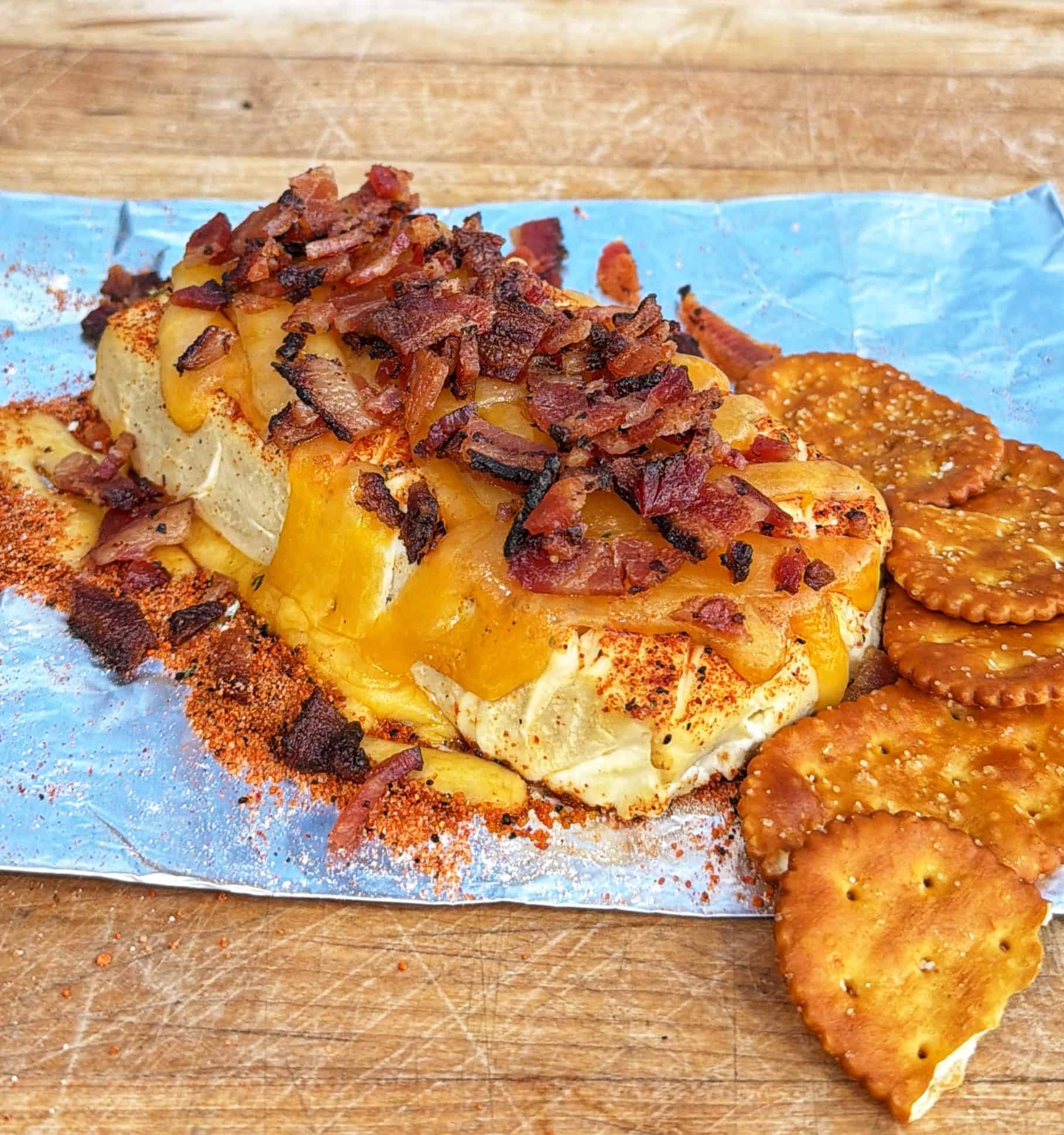 A block of melted cheese topped with crispy bacon bits sits on foil, sprinkled with seasoning, and is served with round crackers on a wooden surface.