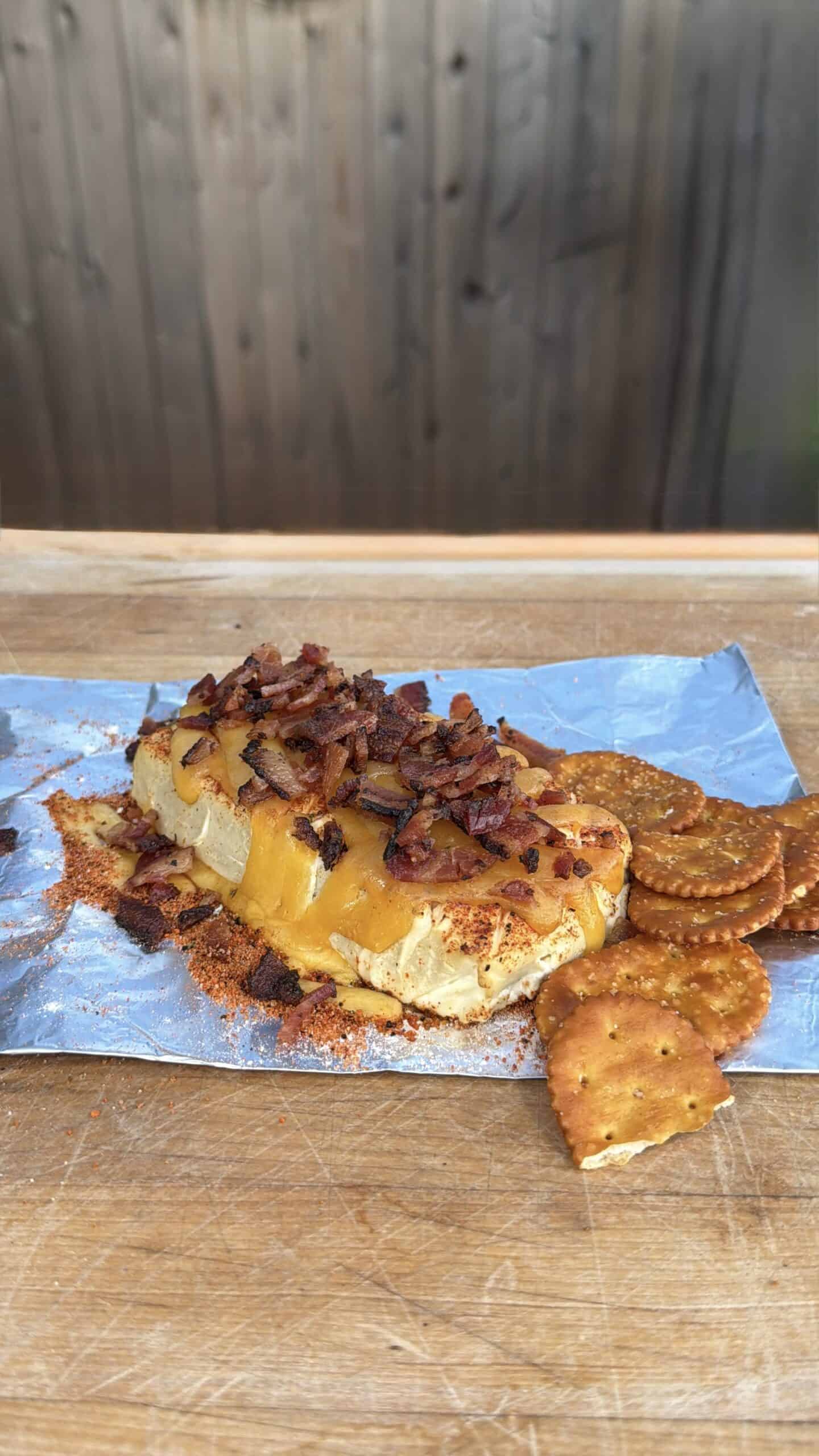 A cheese log topped with crumbled bacon sits on foil, surrounded by round crackers on a wooden surface, with a blurred wooden fence in the background.