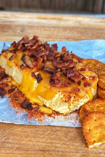 A block of cheese topped with crispy bacon bits and seasoning sits on foil, accompanied by round crackers on a wooden surface.