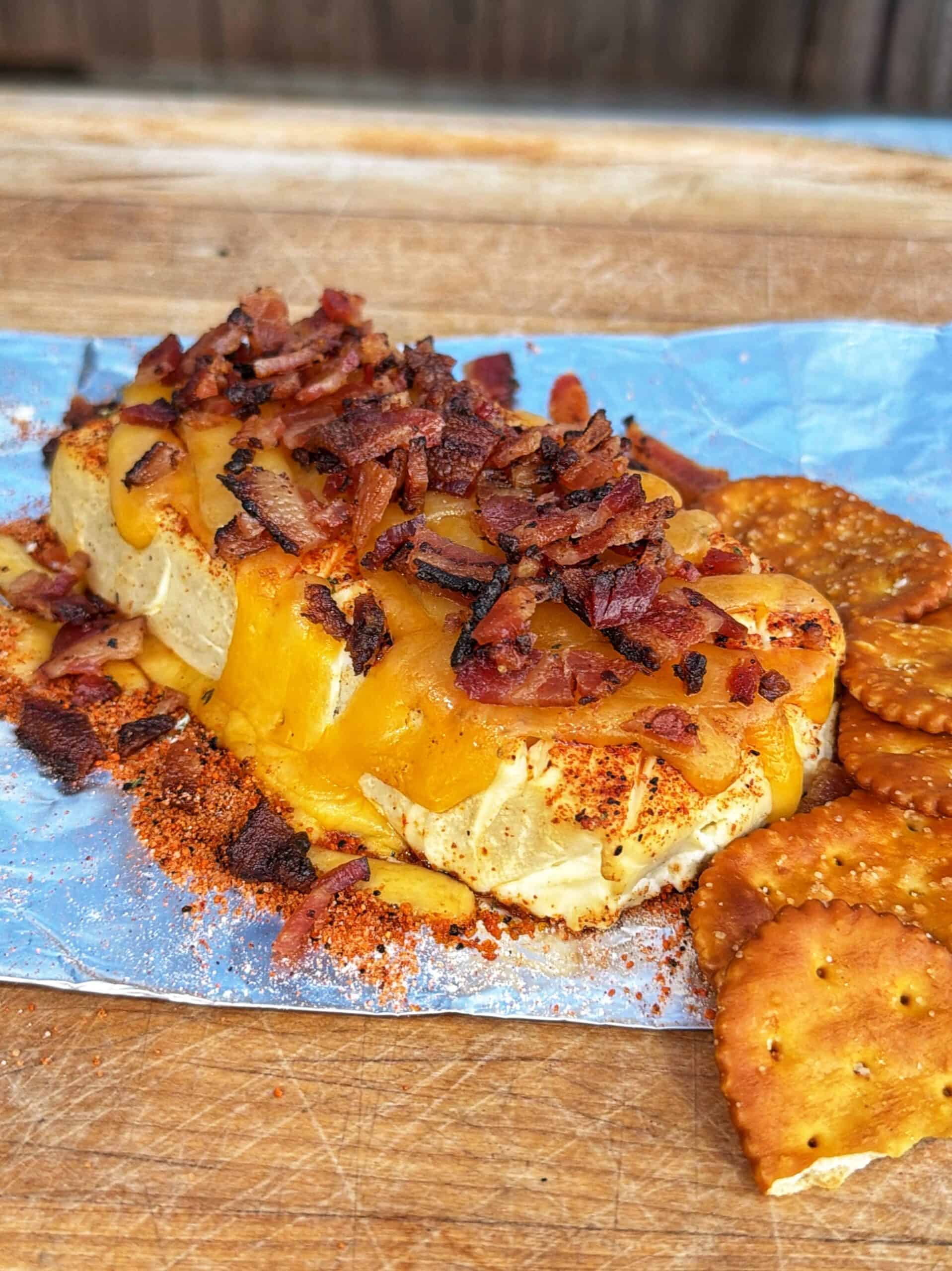 A block of cheese topped with crispy bacon bits and seasoning sits on foil, accompanied by round crackers on a wooden surface.