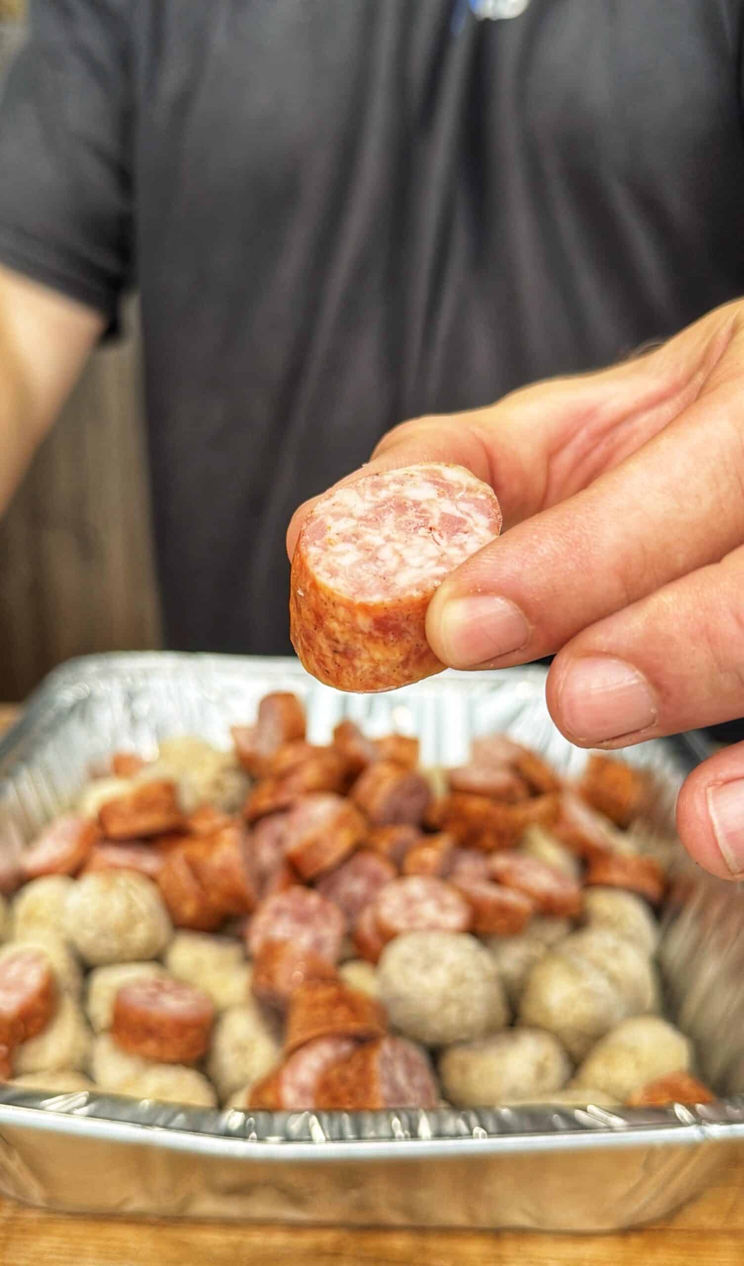 A person holds up a sliced piece of sausage over a tray filled with various sausage slices and pieces, with the focus on the hand and the sausage.