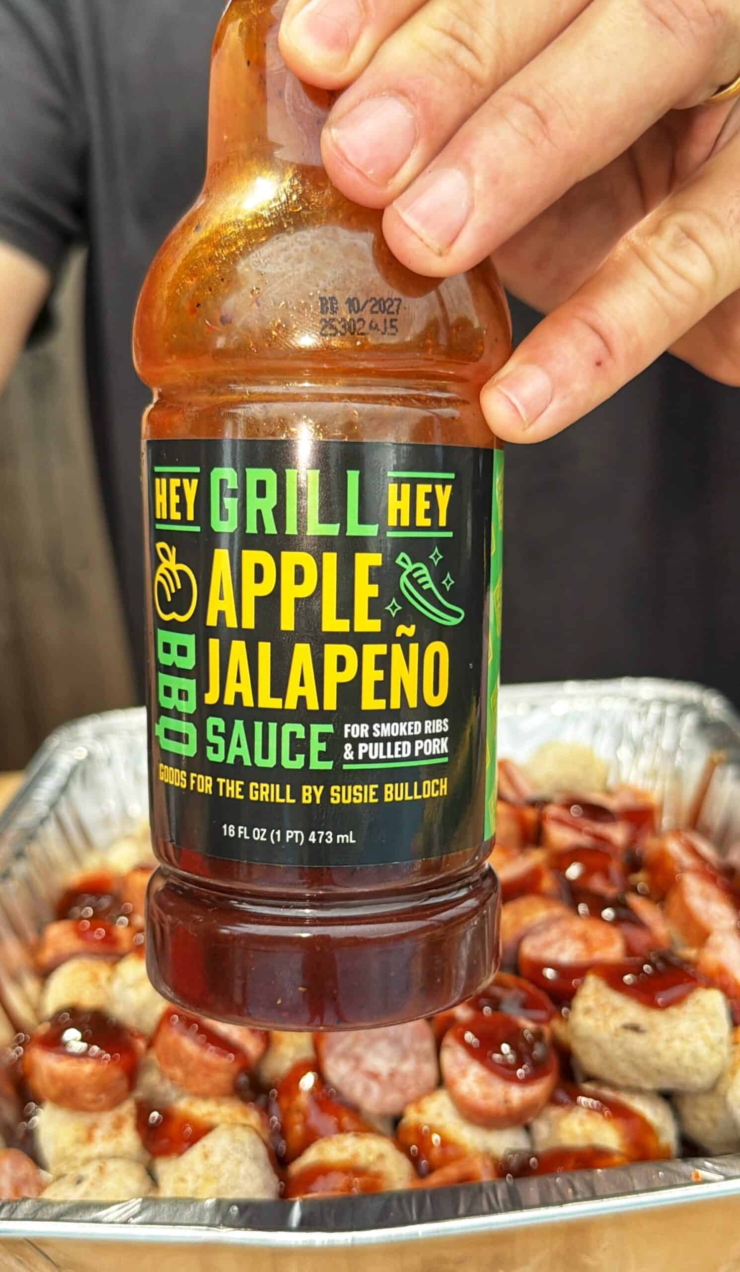 A person holds a bottle of Hey Grill Hey Apple Jalapeño BBQ Sauce over a tray of meat covered in barbecue sauce. The bottle label suggests it’s for smoked ribs and pulled pork.