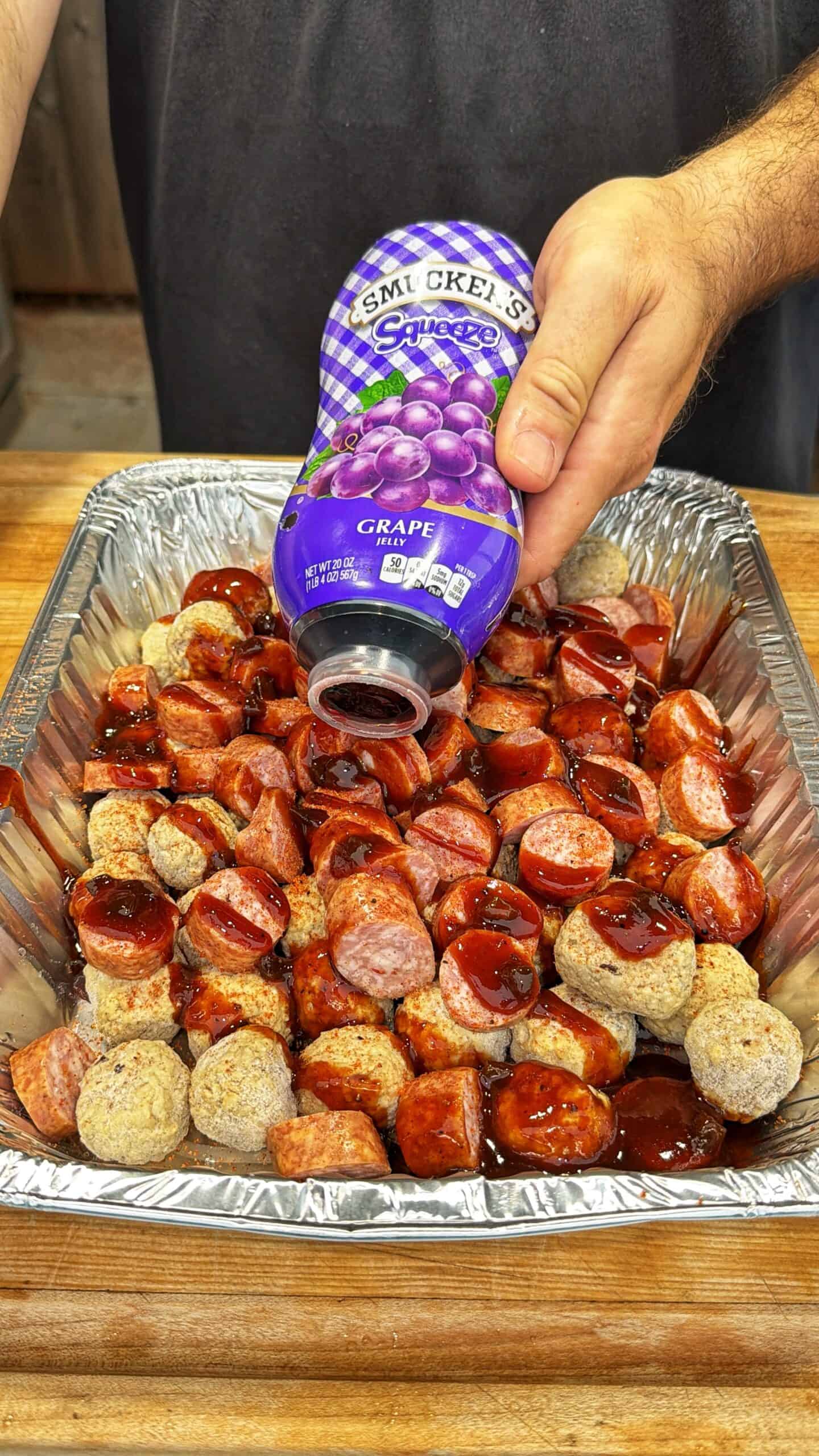 A person pours Smucker’s grape jelly over sliced sausage and meatballs in a large aluminum tray on a wooden surface.