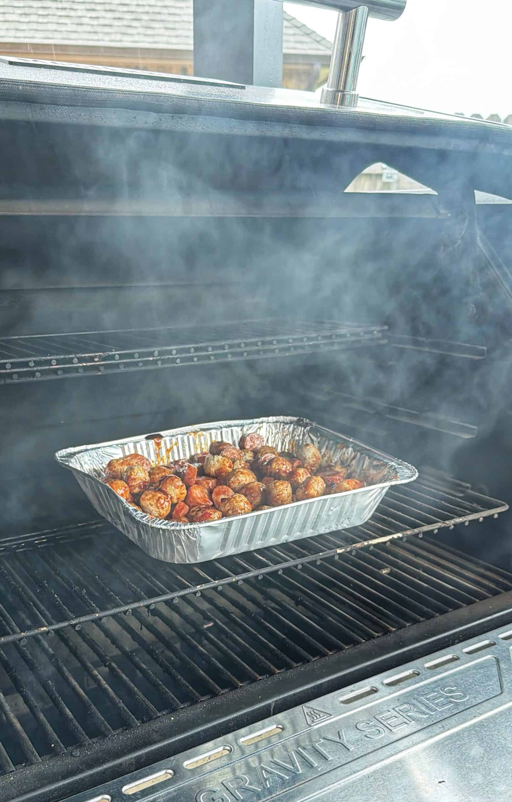 A disposable aluminum tray filled with cooked meatballs sits on a smoker grill, surrounded by smoke. The grill lid is open, and the meatballs appear to be seasoned and browned.