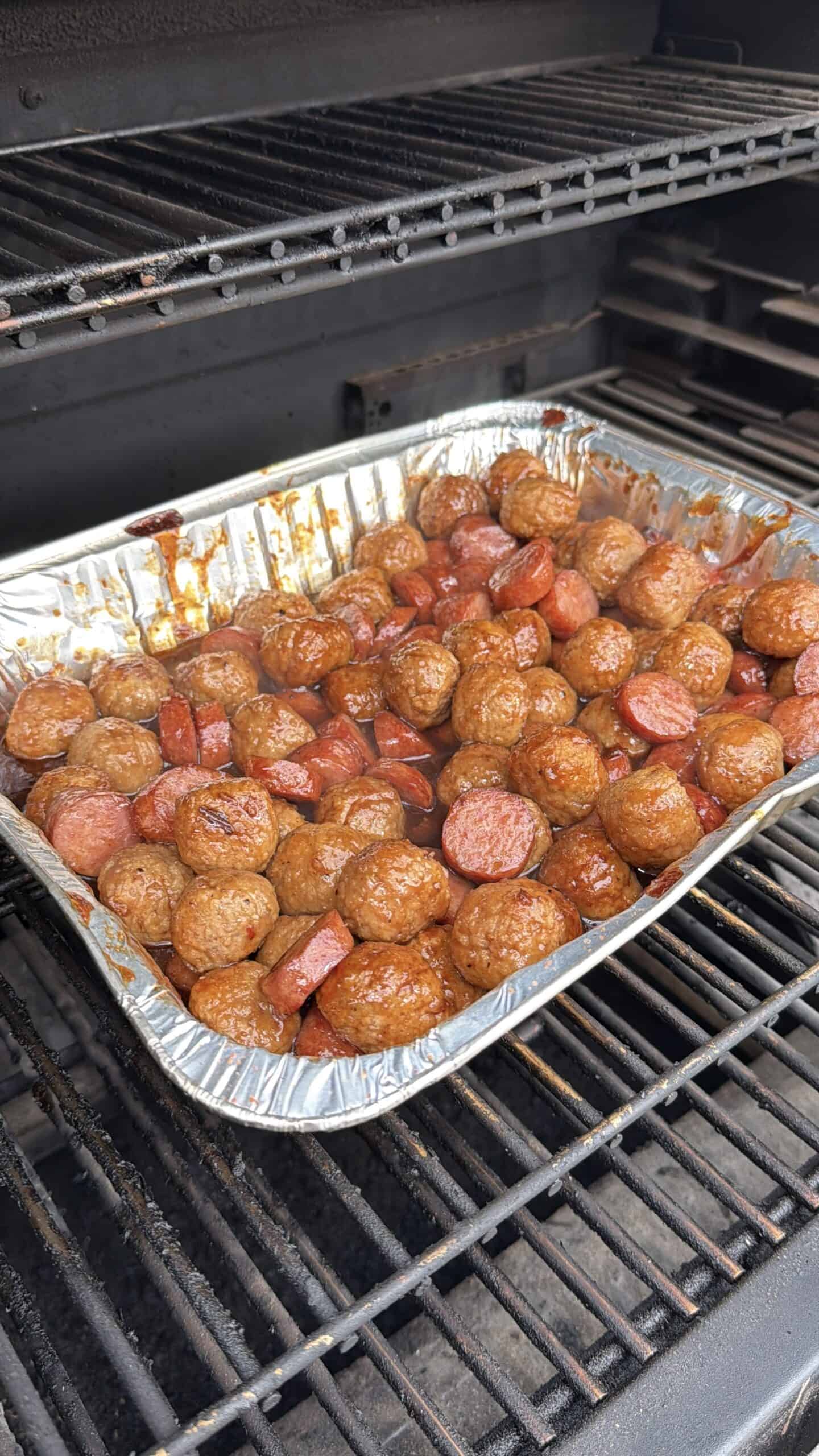 A foil tray filled with meatballs and sliced sausage in sauce sits on the rack inside a barbecue grill, ready to be cooked.