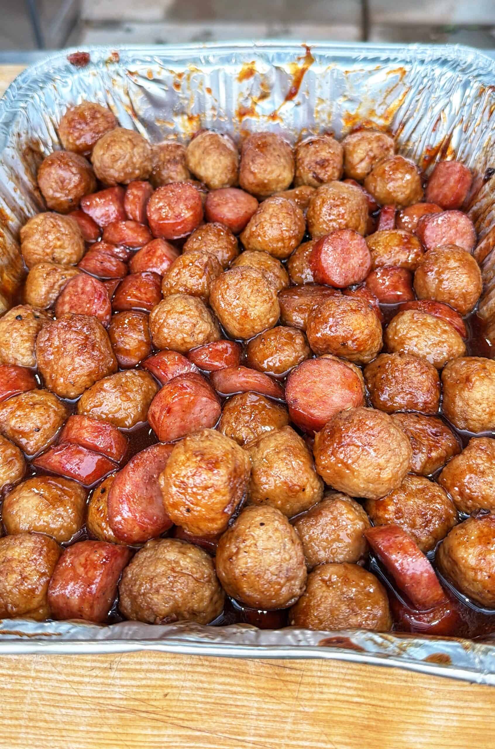 A foil tray filled with glazed meatballs and sliced sausage pieces, all coated in a rich brown sauce, sitting on a wooden surface.