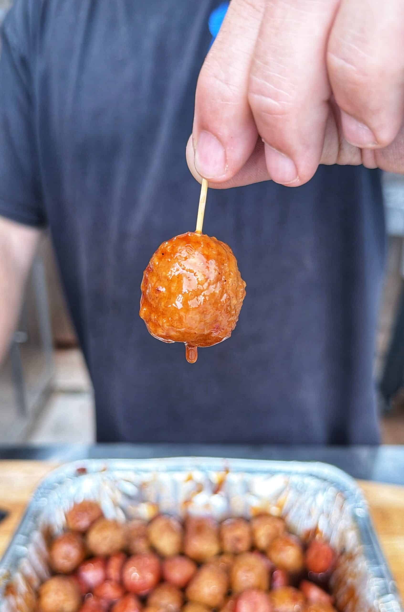 A hand holds a toothpick with a glazed meatball above a tray filled with more meatballs.
