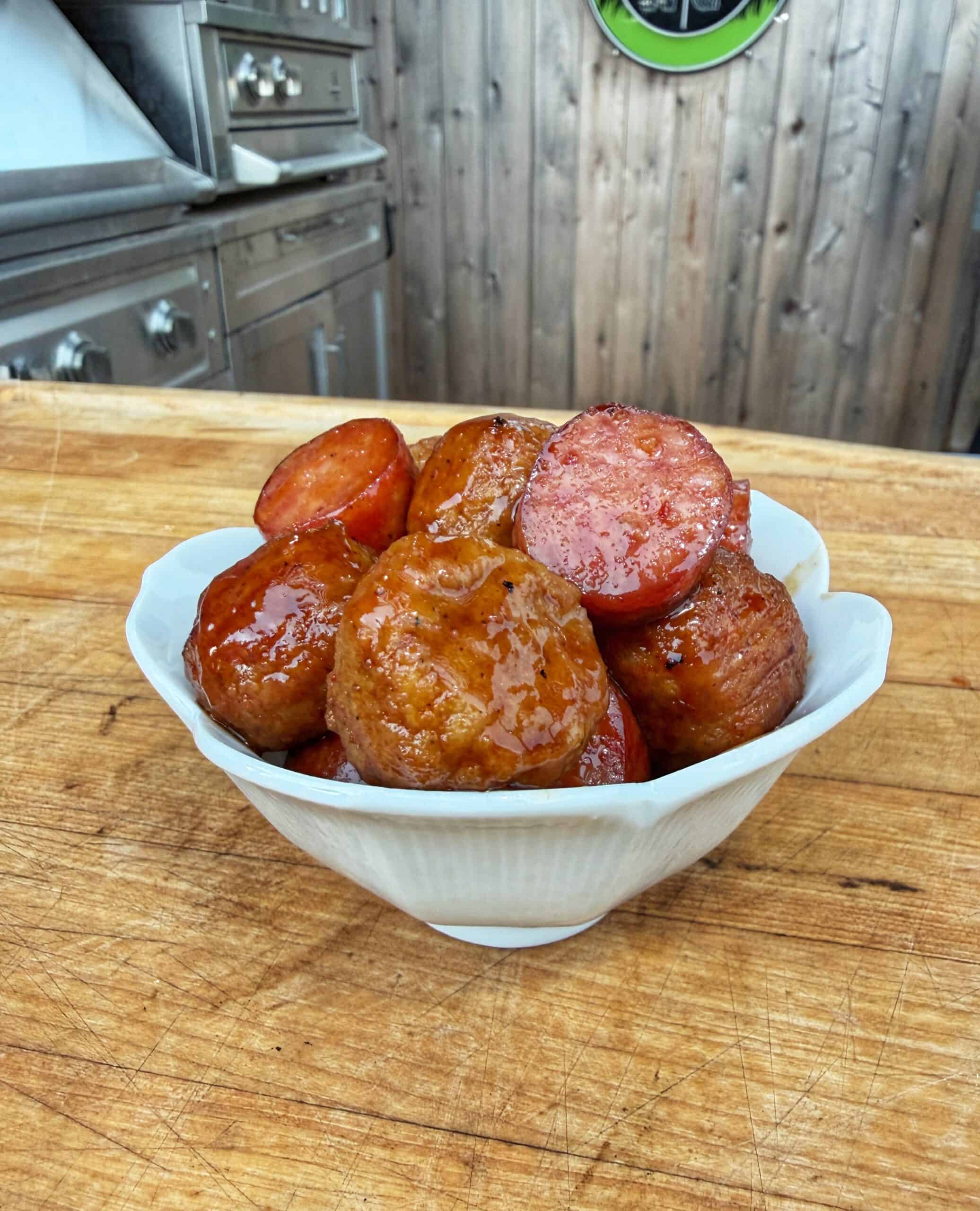 A white bowl filled with glazed, round pieces of smoked sausage and meat, placed on a wooden counter with an outdoor grill and wooden wall in the background.