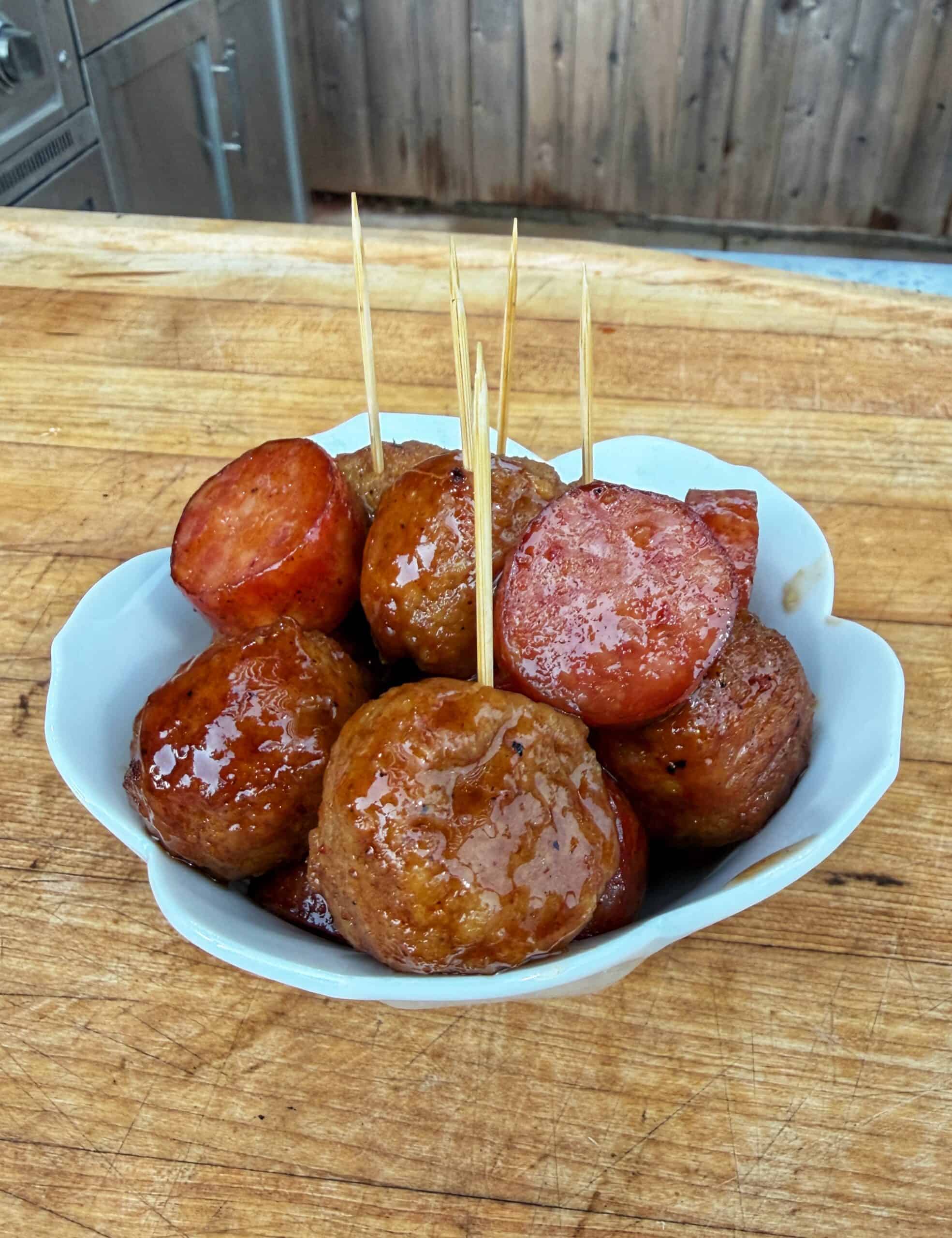 A white bowl filled with glazed meatballs and sliced sausage, each with a wooden toothpick, sits on a wooden surface. The food appears shiny and saucy, ready to be served as an appetizer.