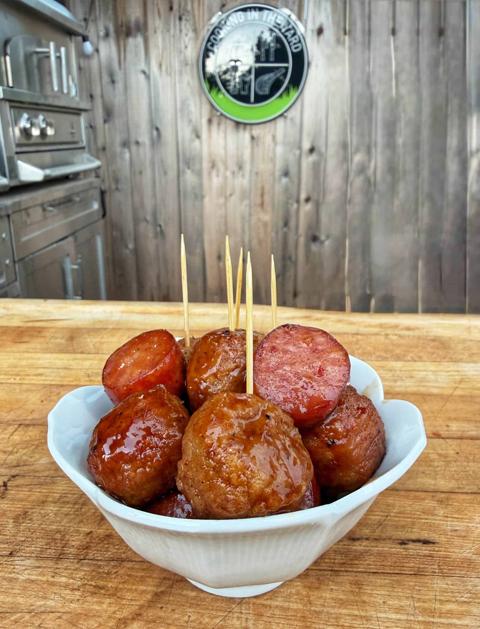 A white bowl filled with glazed meatballs and slices of sausage, each with a toothpick, sits on a wooden counter. In the background, theres an outdoor grill and a wooden fence with a round grilling sign.