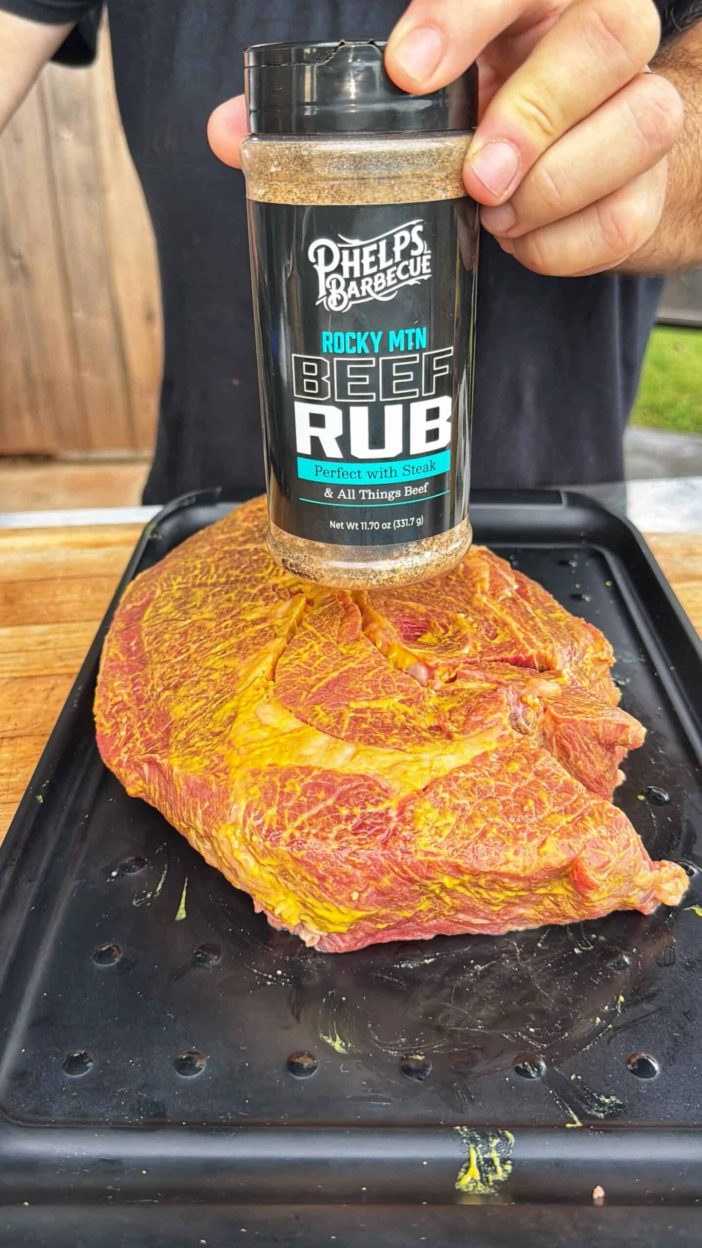 A hand holds a container of Phelps Barbecue Rocky MTN Beef Rub over a large, seasoned cut of raw beef placed on a black tray, ready for grilling or smoking outdoors.