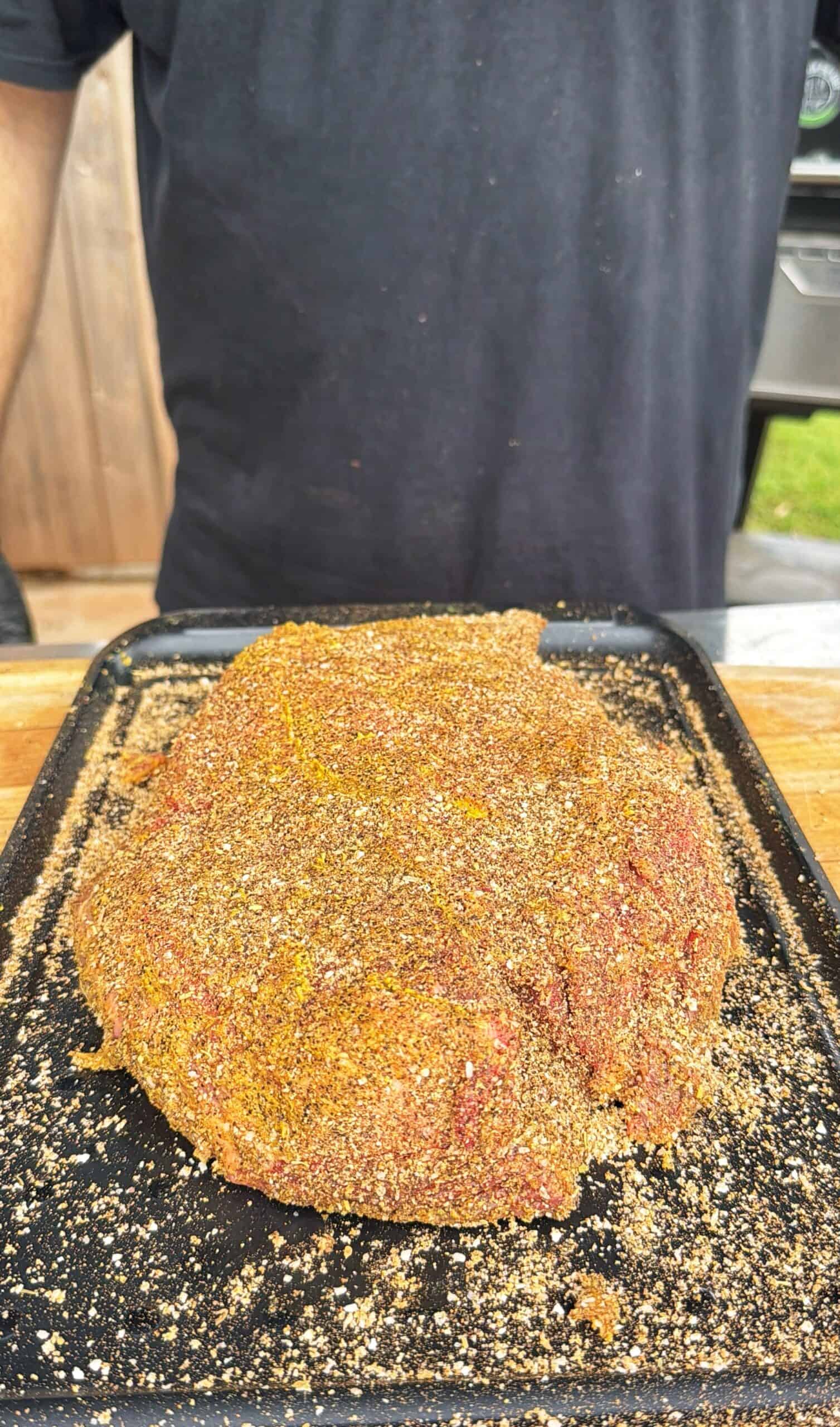 A large piece of raw meat coated in seasoning sits on a black tray, with a person in a black shirt standing behind it.