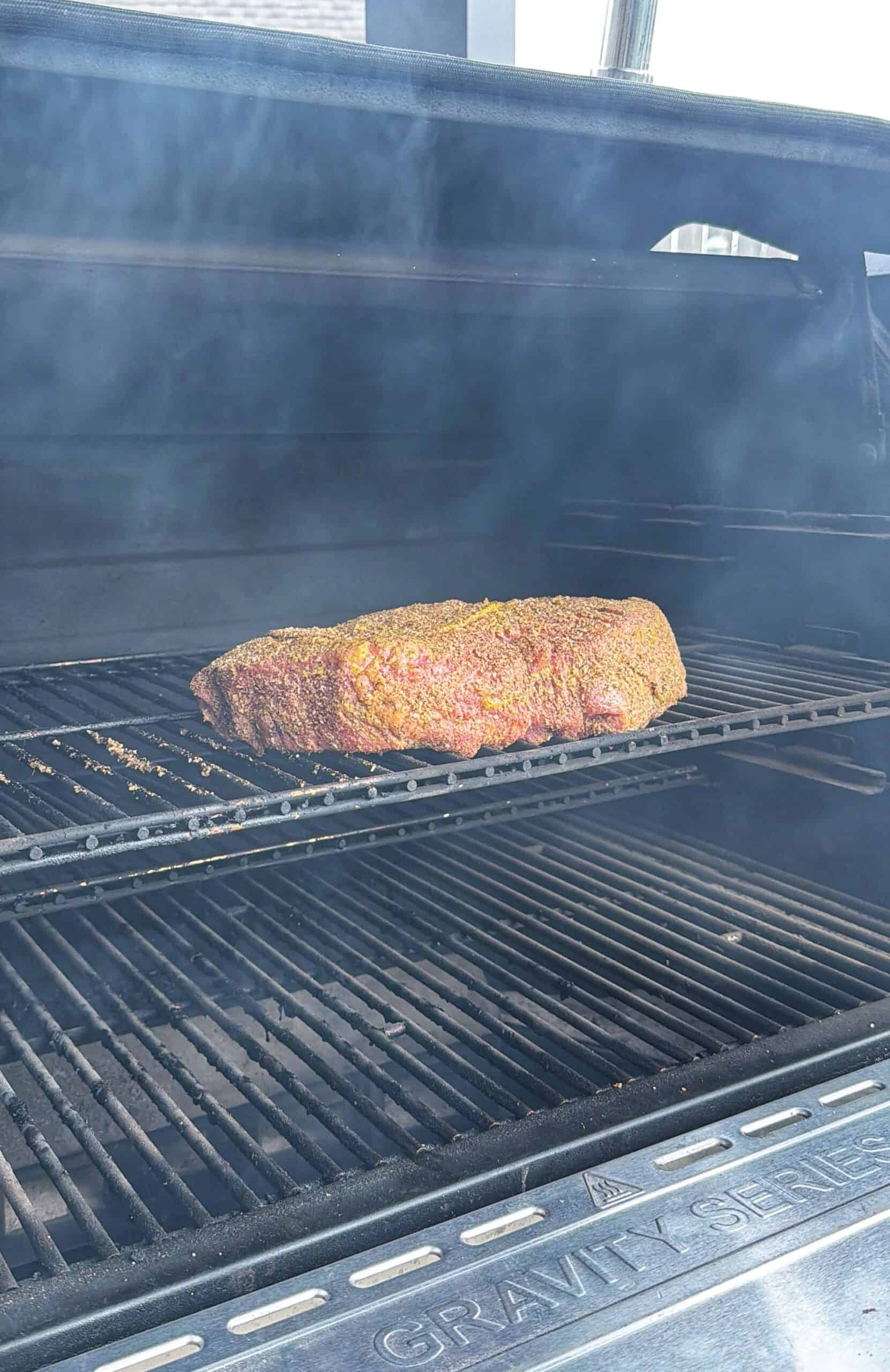 A seasoned brisket cooks on the upper rack of a smoky outdoor grill, with visible smoke and open grill lid, as sunlight shines on the meat.