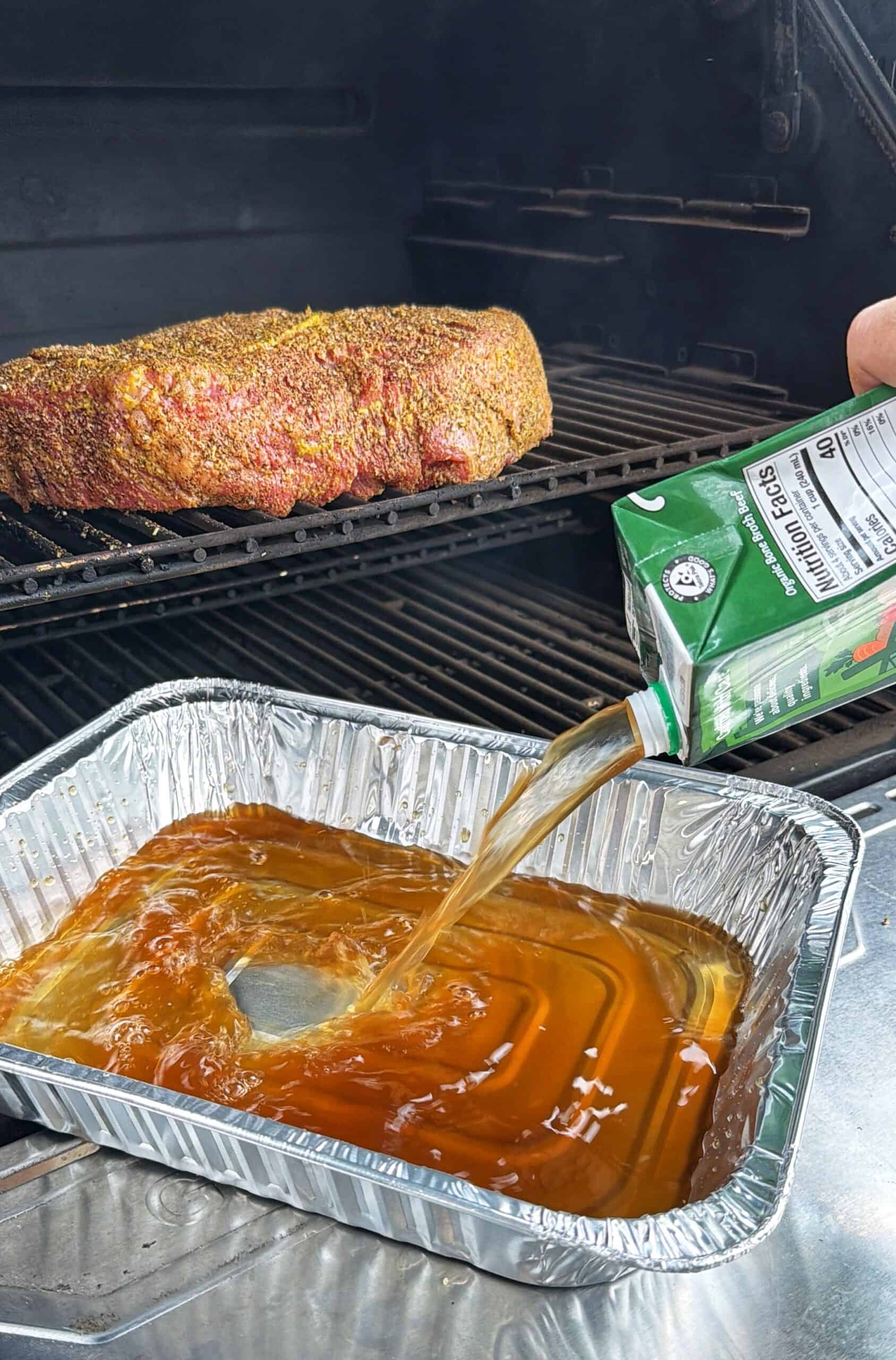 A person pours apple juice from a carton into a foil pan on a grill, while a seasoned piece of meat cooks on the upper rack above the pan.