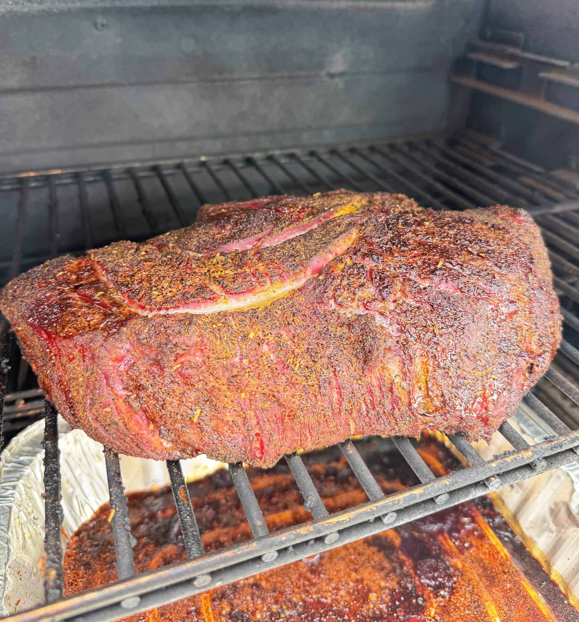 A large, seasoned brisket sits on the top rack of a smoker, developing a dark, flavorful crust as it cooks. A <a href=