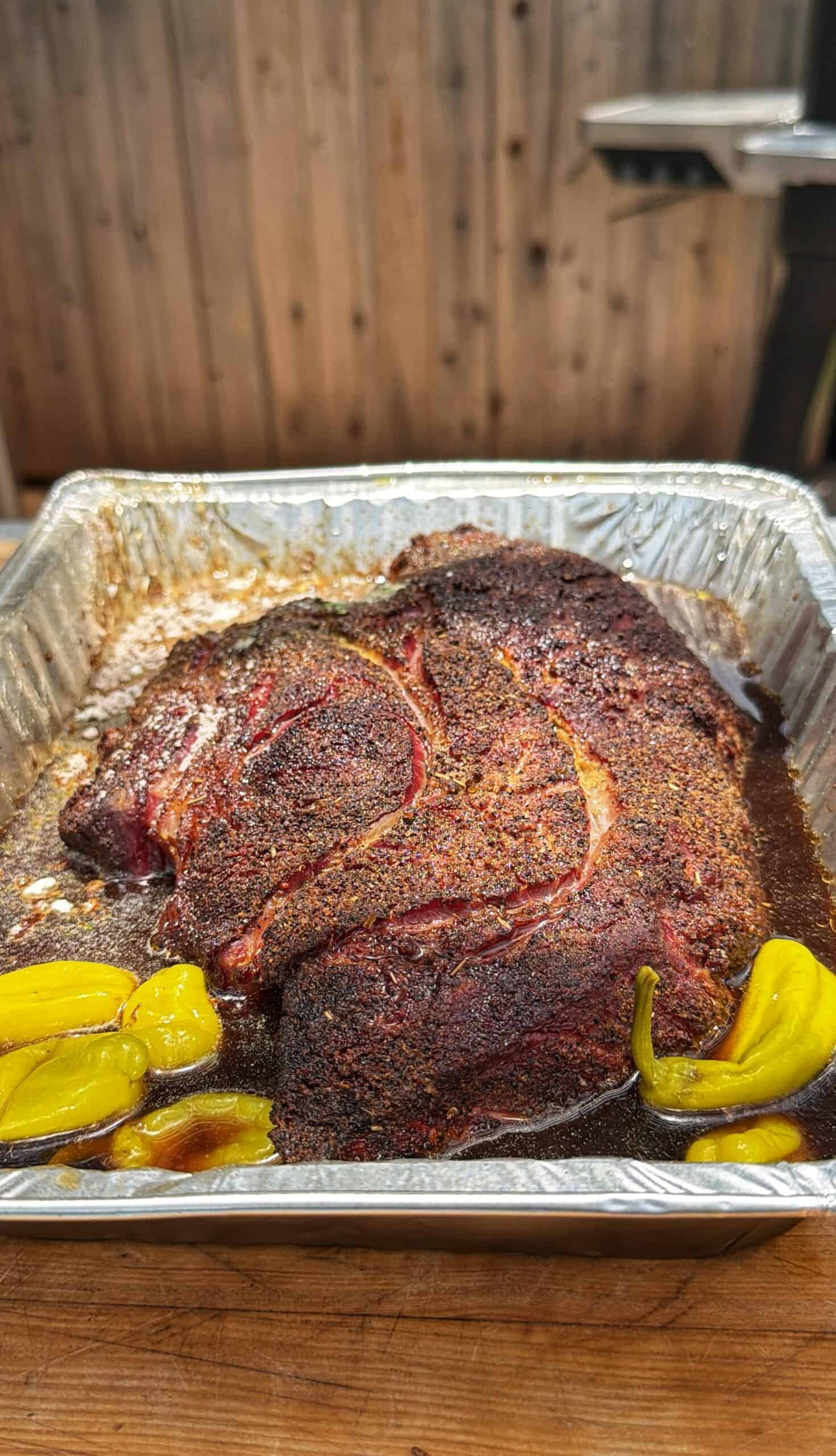 A seasoned, cooked beef roast sits in a foil tray with juices and a few yellow peppers, set on a wooden surface with a wooden fence and grill in the blurry background.