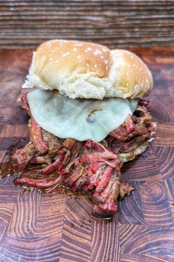 A pulled barbecue beef sandwich with melted cheese on a sesame seed bun, sitting on a wooden surface with a rustic background.