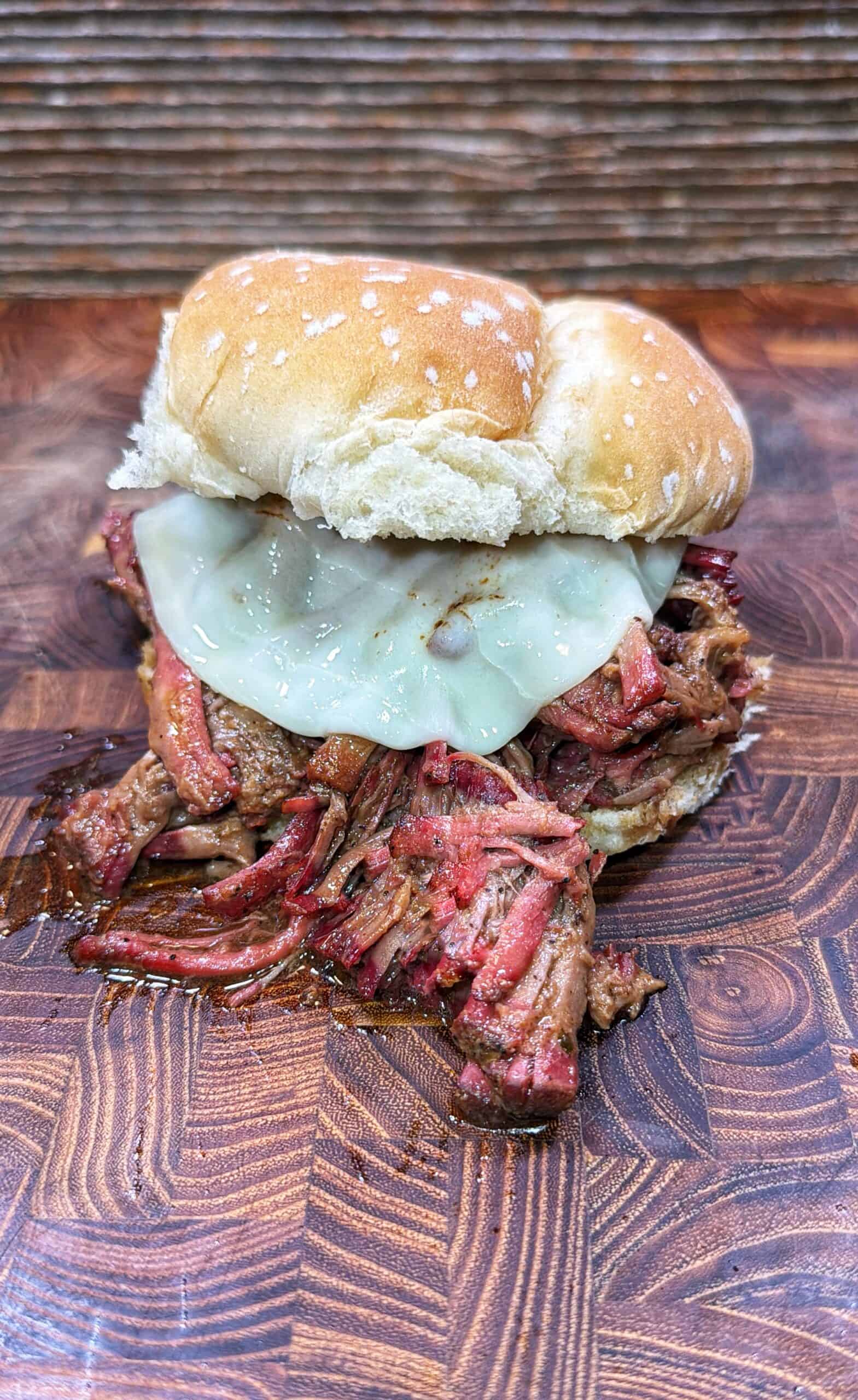 A pulled barbecue beef sandwich with melted cheese on a sesame seed bun, sitting on a wooden surface with a rustic background.