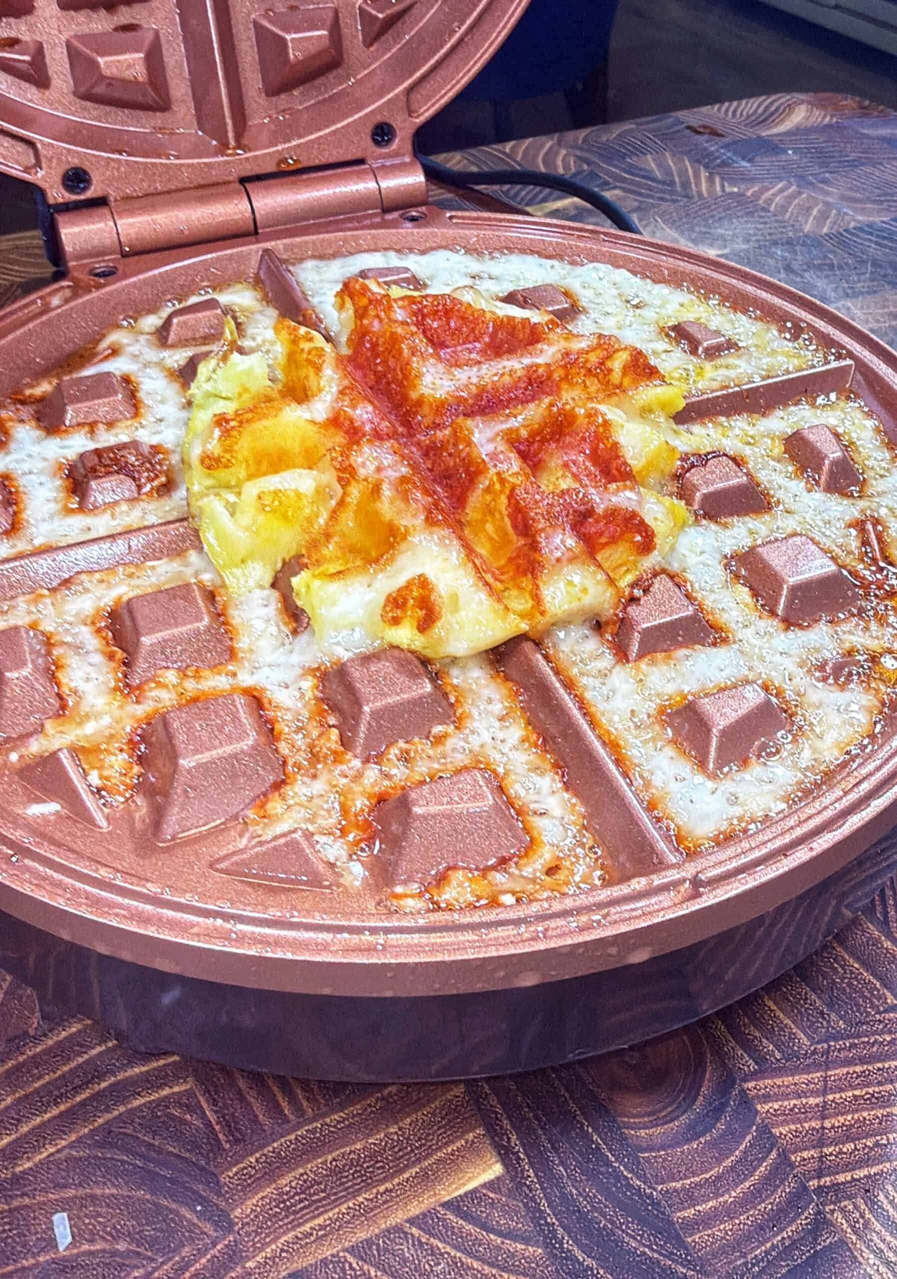 A round waffle maker with an open lid contains a partly cooked, melting mixture of cheese and sauce forming a waffle shape, with some cheese spilling over onto the hot surface.